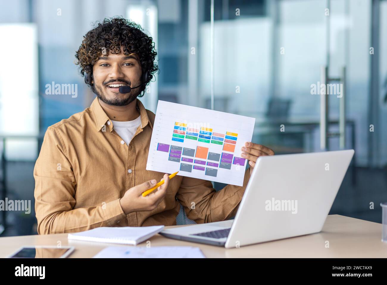 Uomo professionista sorridente che organizza una riunione virtuale, che presenta una colorata mappa di Gantt, con cuffie e laptop in un ufficio moderno. Foto Stock