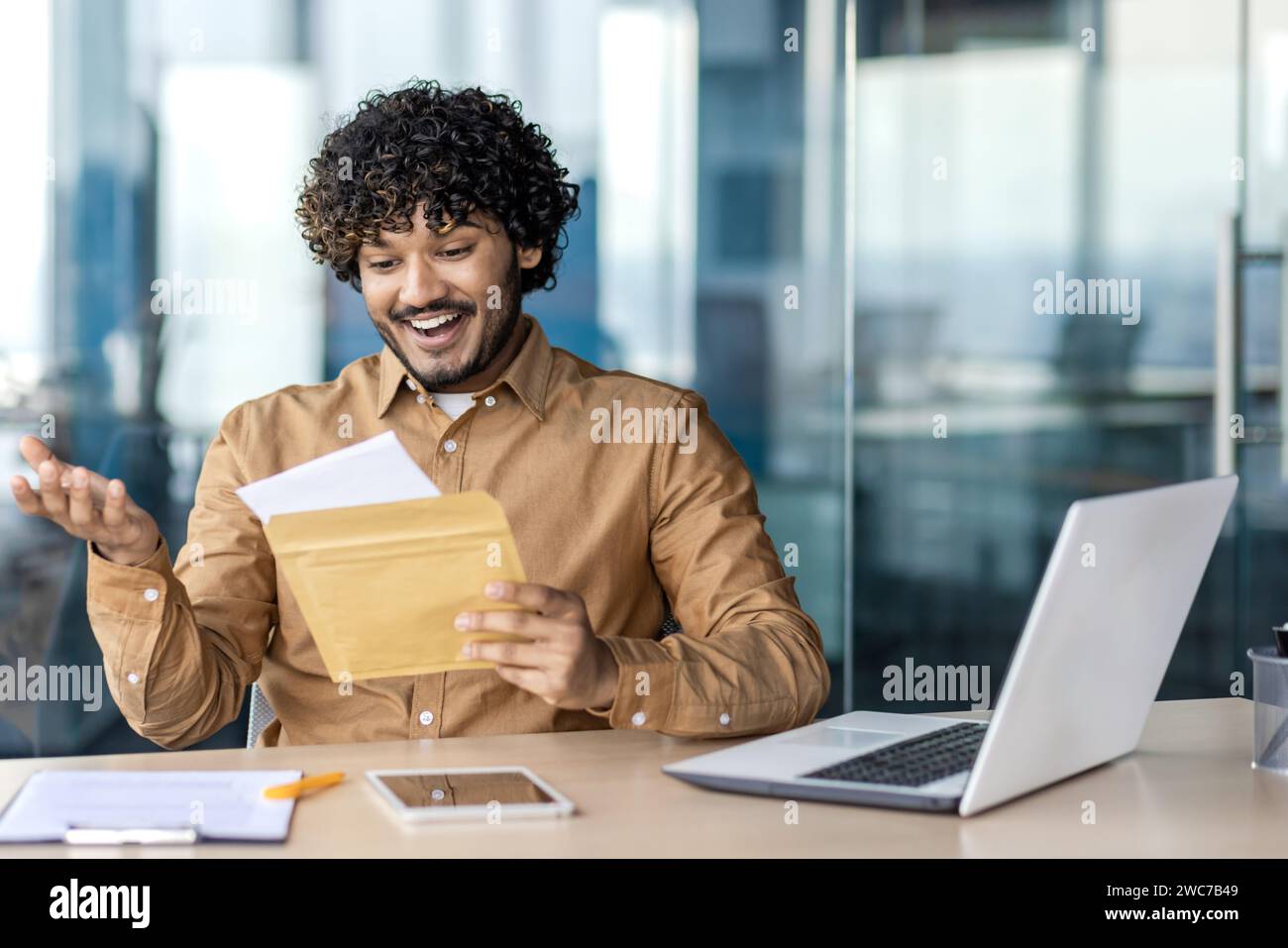 Un giovane eccitato in un abbigliamento casual da lavoro reagisce con gioia all'apertura di un pacchetto alla sua scrivania. Foto Stock