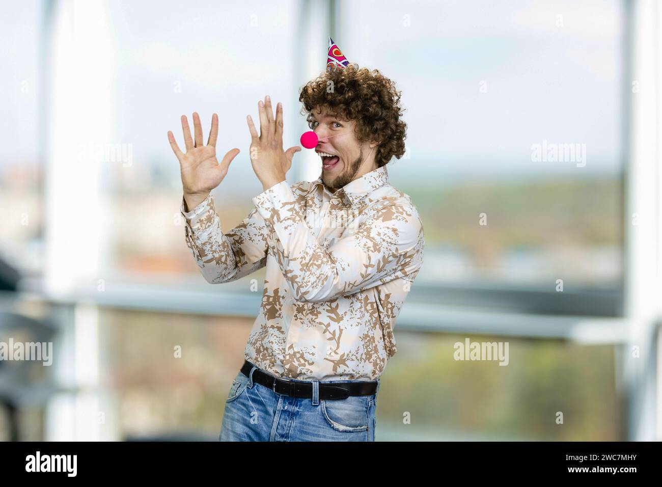 Felice ragazzo caucasico con i capelli ricci che indossa un naso di clown. Finestra interna sullo sfondo. Foto Stock