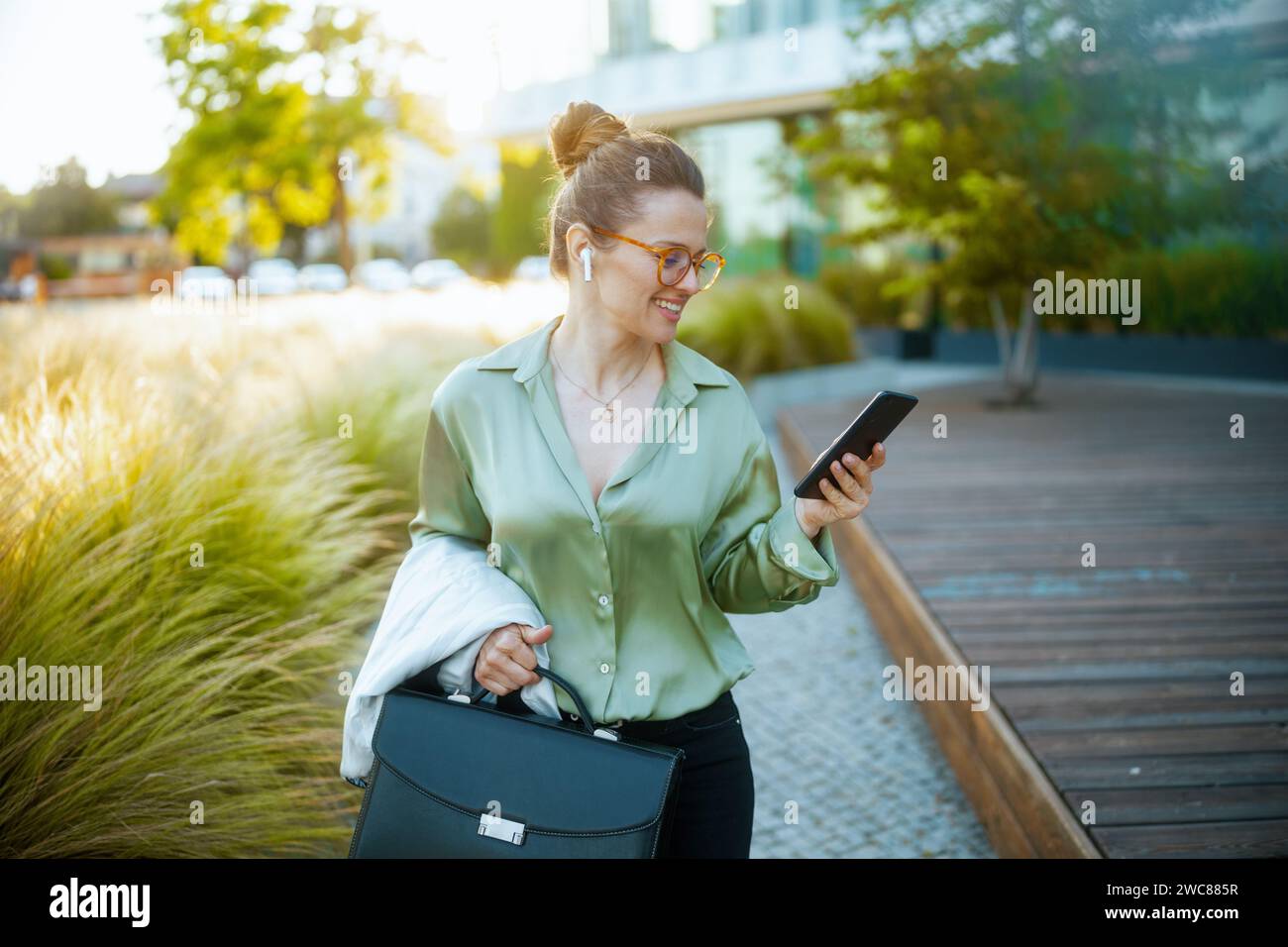 donna moderna sorridente lavoratrice vicino al business center con camicia verde e occhiali da vista con valigetta e cuffie wireless tramite smartphone. Foto Stock