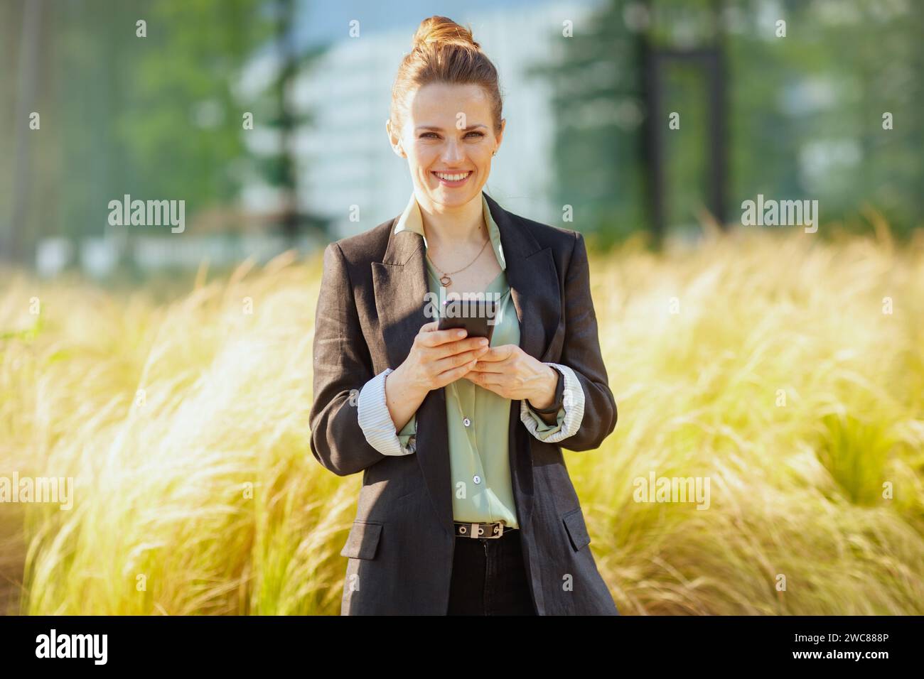 donna d'affari moderna e felice di mezza età nel quartiere degli affari con giacca nera e smartphone. Foto Stock