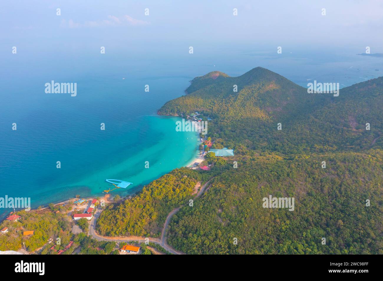 Isola tropicale con una baia di sabbia azzurra e un molo e un villaggio sulla spiaggia circondato da strade da foreste esotiche, alberi e colline, vista aerea. Foto Stock
