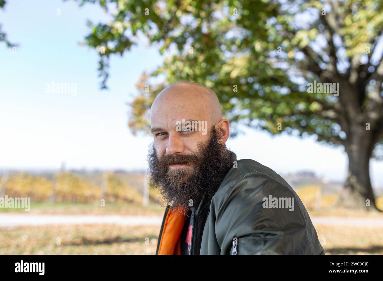 Un uomo con una barba lunga e incasinata sorridente con il sole. È all'aperto, calvo e indossa una giacca da bombardamento Foto Stock