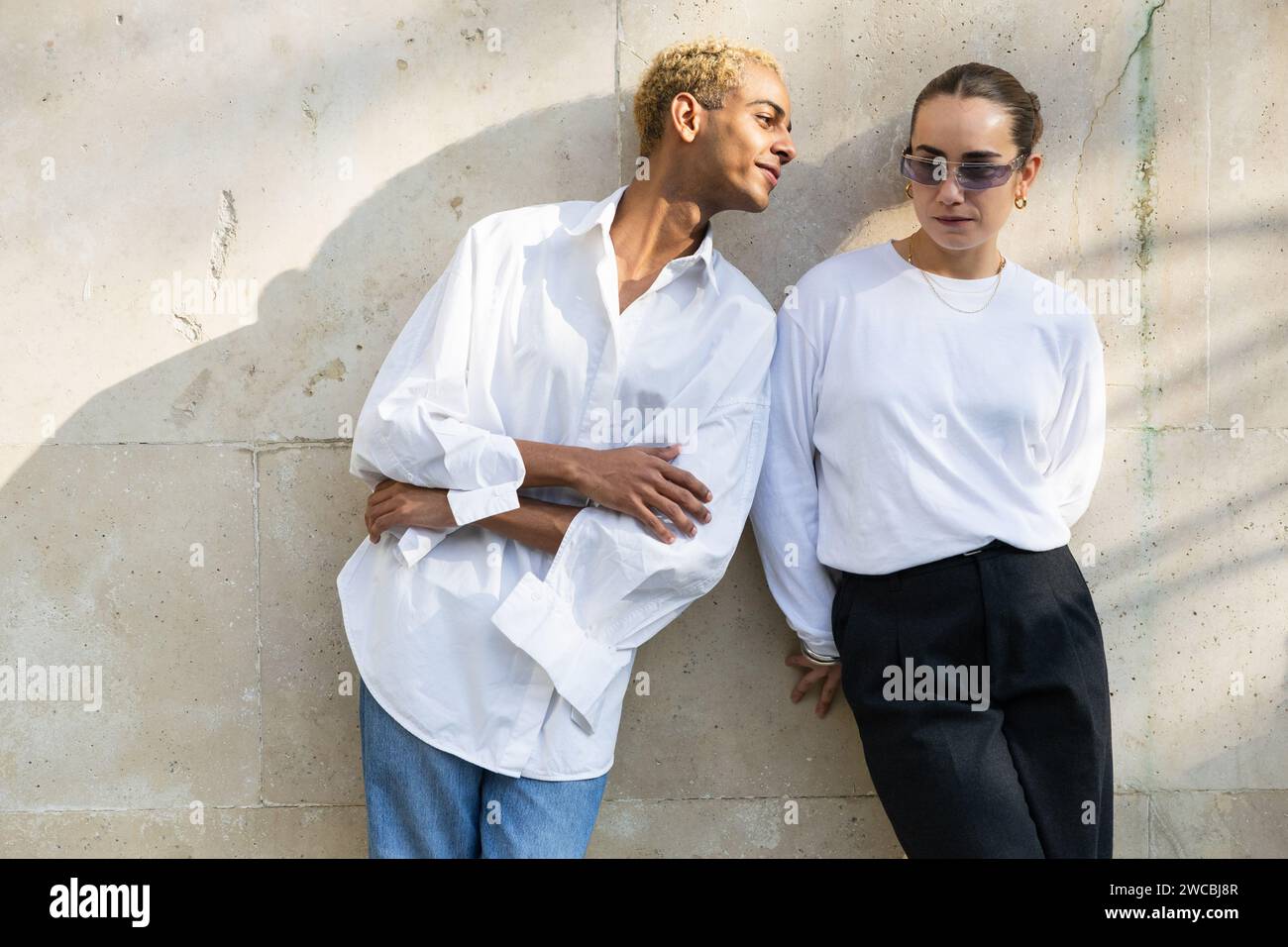 Un giovane uomo e una donna in elegante stile minimalista si appoggiano a una parete di cemento, trasudando un'atmosfera urbana di tendenza. Foto Stock