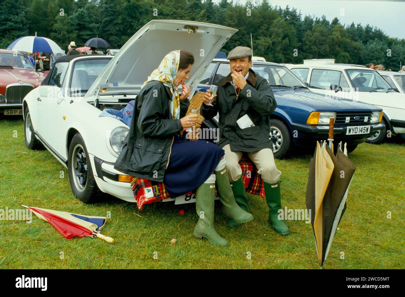 La coppia Sloane Ranger si sta godendo un picnic all'aperto dalla Porsche 911 SC Cabriolet decappottabile. E' su un Bucks Fizz, ma fatto con Henkell Trocken, un vino bianco frizzante tedesco, mentre lui lo sta prendendo in ordine. Entrambi sono vestiti come dettano la moda per l'occasione con nuovi stivali Hunter Wellington verdi senza fango e le sue giacche blu cerate Burberry, assolutamente in voga all'epoca. Ha indossato un velo Hermes, mentre lui è in un cappellino piatto tweed. Windsor, Berkshire, Inghilterra giugno 1985 1980s HOMER SYKES Foto Stock