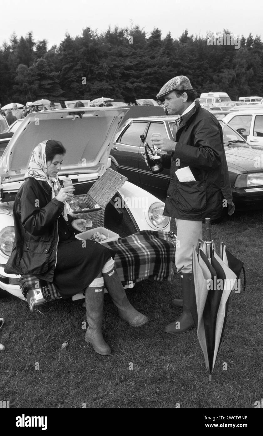 Polo Windsor Great Park, presso il Guards Polo Club Ground, una coppia di Sloane Ranger alla moda che si gode un picnic all'aperto dalla loro Porsche 911 SC Cabriolet decappottabile. E' su un Bucks Fizz, ma fatto con Henkell Trocken, un vino bianco frizzante tedesco, mentre lui lo sta prendendo in ordine. Entrambi sono vestiti come dettano la moda per l'occasione con nuovi stivali Hunter Wellington verdi senza fango e le sue giacche blu cerate Burberry, assolutamente in voga all'epoca. Ha indossato un velo Hermes, mentre lui è in un cappellino piatto tweed. Windsor, Berkshire, Inghilterra giugno 1985 1980s HOMER SYKES Foto Stock
