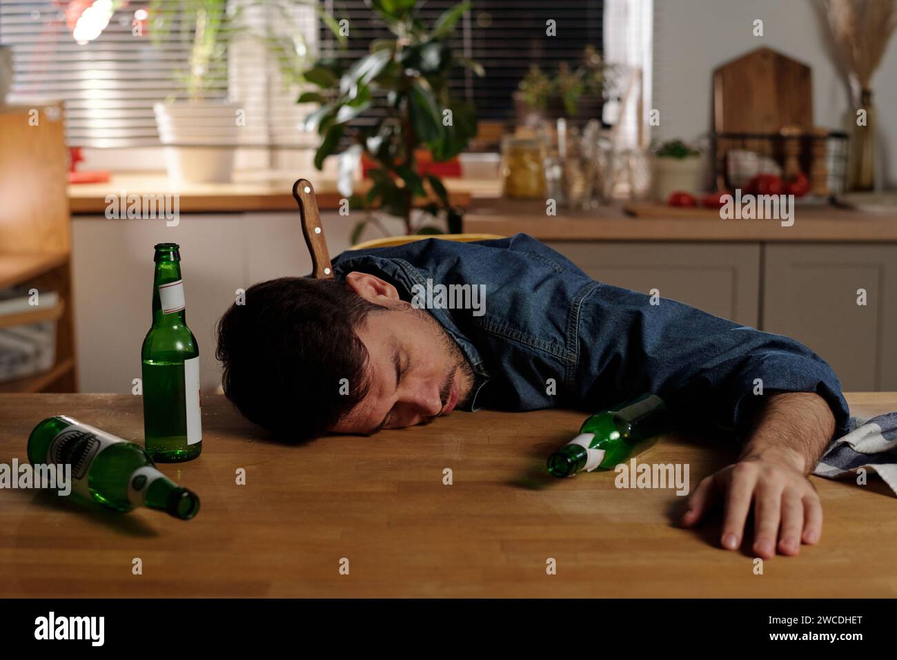 Giovane uomo depresso dipendente dall'alcol con un coltello nel collo che dorme sul tavolo della cucina con diverse bottiglie di birra vuote Foto Stock