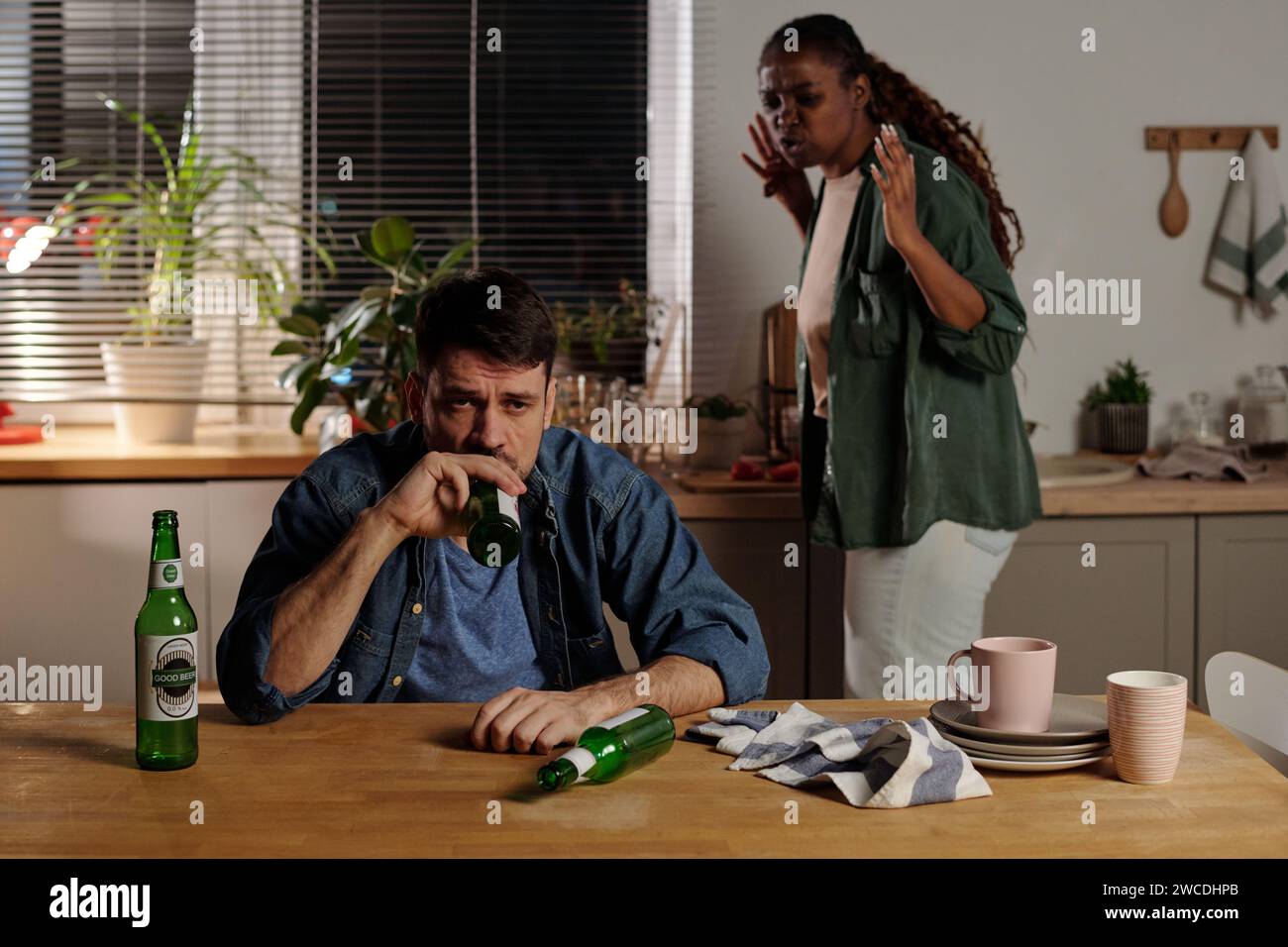 Uomo infelice con dipendenza da alcol seduto a tavola in cucina e bevendo birra contro moglie irritata che urla contro di lui durante il conflitto Foto Stock