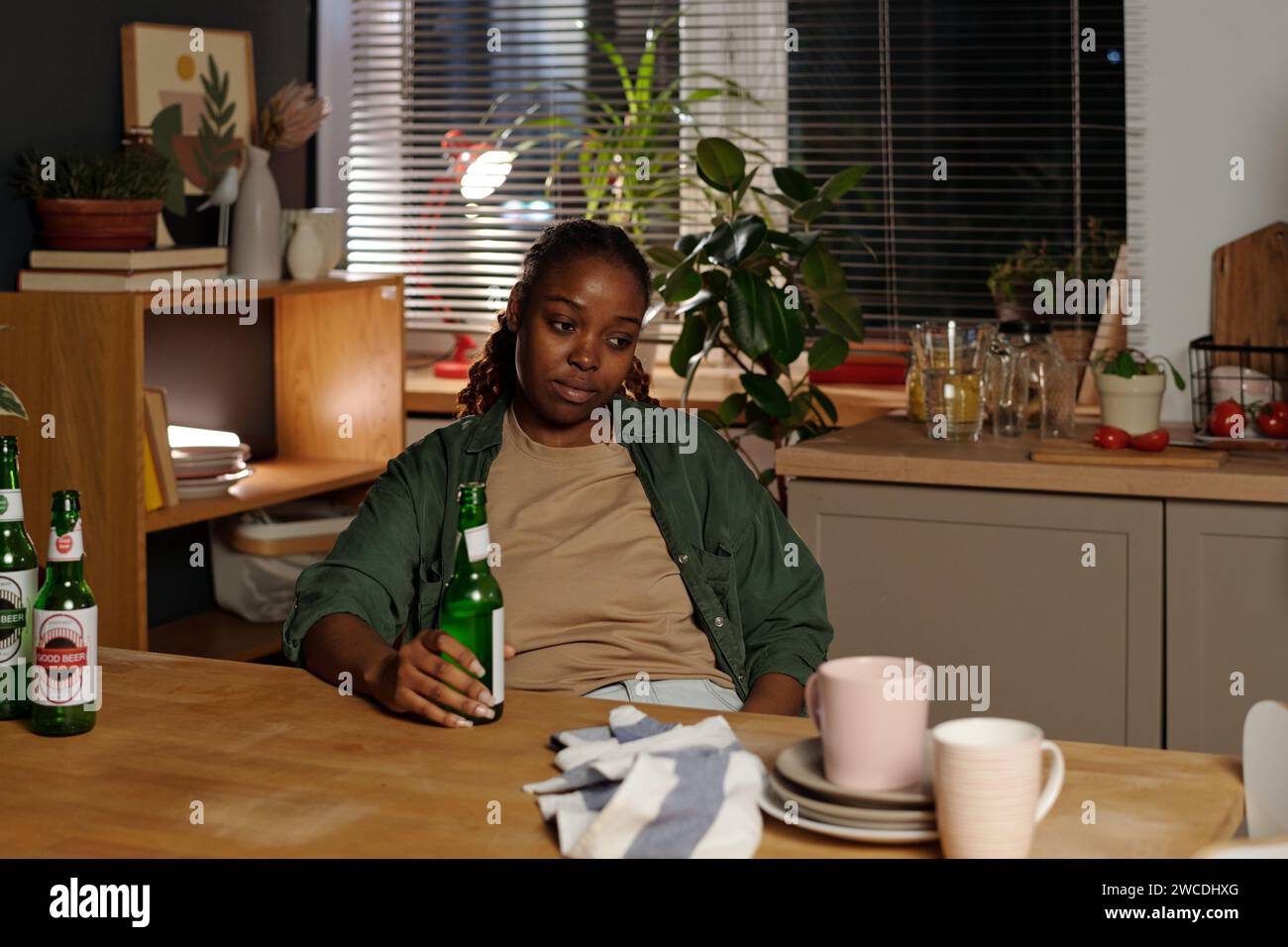 Giovane donna afro-americana depressa che soffre di dipendenza alcolica tenendo una bottiglia di birra vuota mentre si siede al tavolo della cucina Foto Stock
