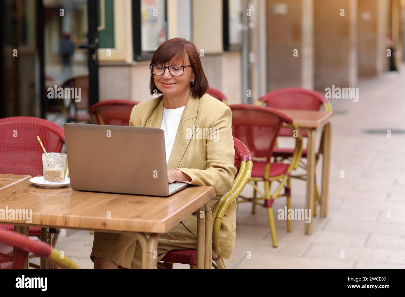Donna d'affari matura ed elegante con occhiali da vista lavora sul computer portatile mentre si siede con un caffè all'aperto in un caffè europeo. Concetto di lavoro remoto da pub Foto Stock