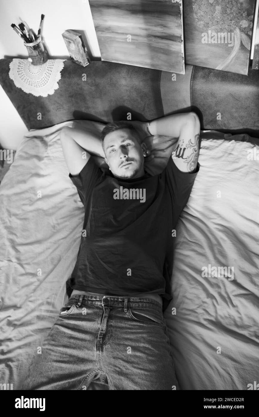Giovane artista che posa sul letto sullo sfondo di dipinti. Bell'uomo con tatuaggi in posa a letto in un ambiente creativo che mostra i dipinti che crea Foto Stock