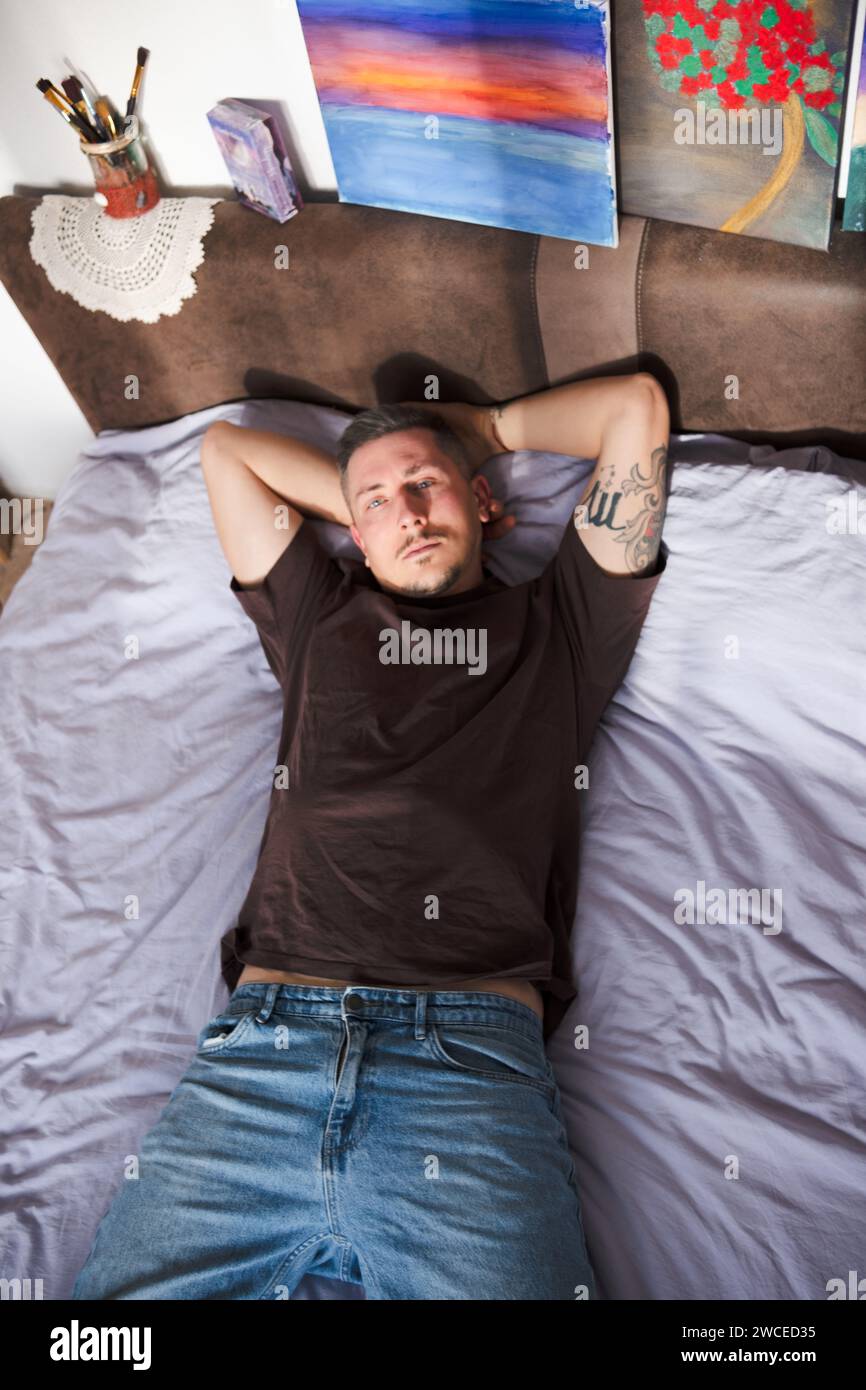 Giovane artista che posa sul letto sullo sfondo di dipinti. Bell'uomo con tatuaggi in posa a letto in un ambiente creativo che mostra i dipinti che crea Foto Stock