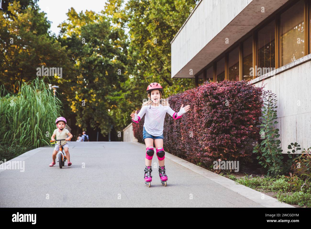 Due bambine cavalcano su rulli e in bici nel parco estivo. Bambini che indossano cuscini di protezione e casco di sicurezza per una guida sicura. Sport all'aperto attivo f Foto Stock
