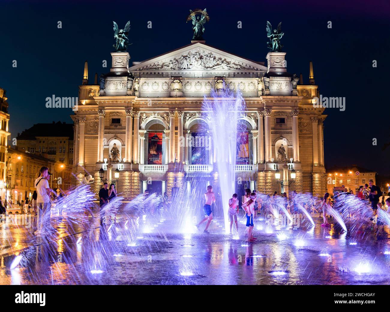 Bambini sotto i ruscelli di una fontana illuminata sulla piazza di fronte all'Opera Nazionale di Leopoli su un veicolo elettrico estivo Foto Stock
