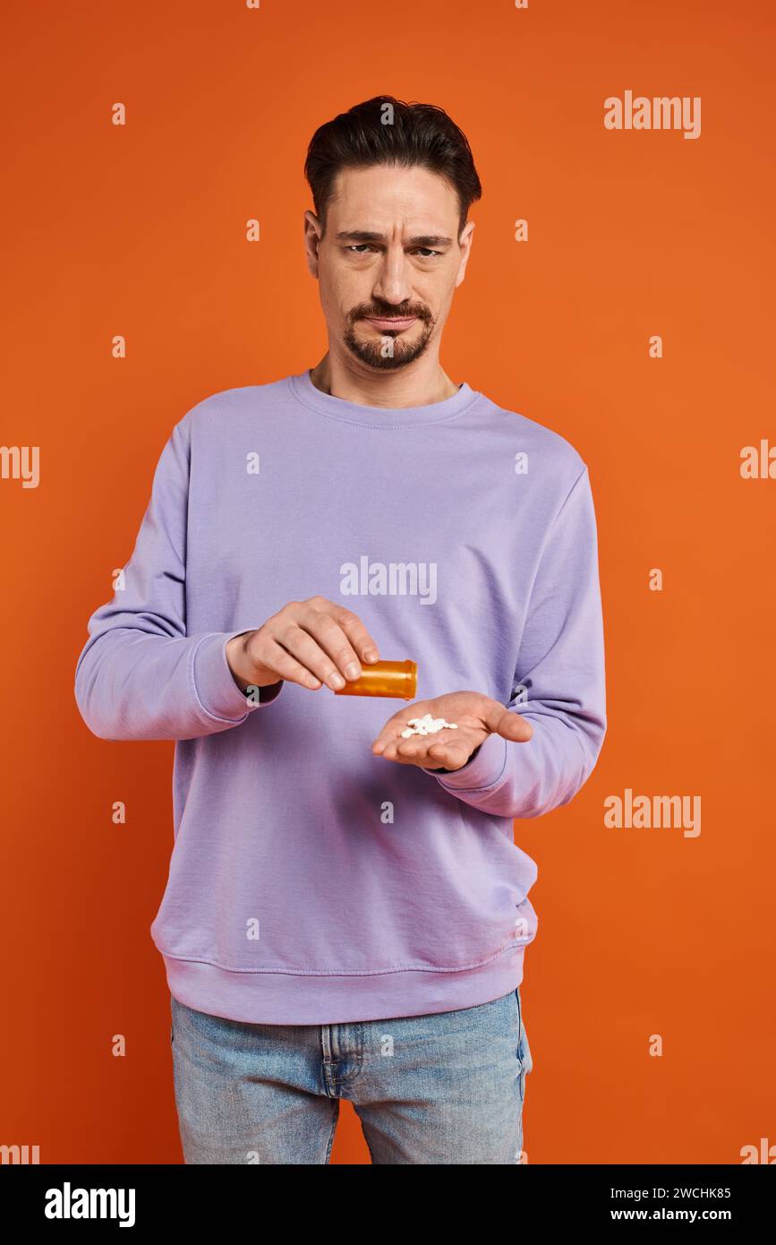 uomo barbuto in felpa viola che versa il farmaco in mano su sfondo arancione, pillole Foto Stock