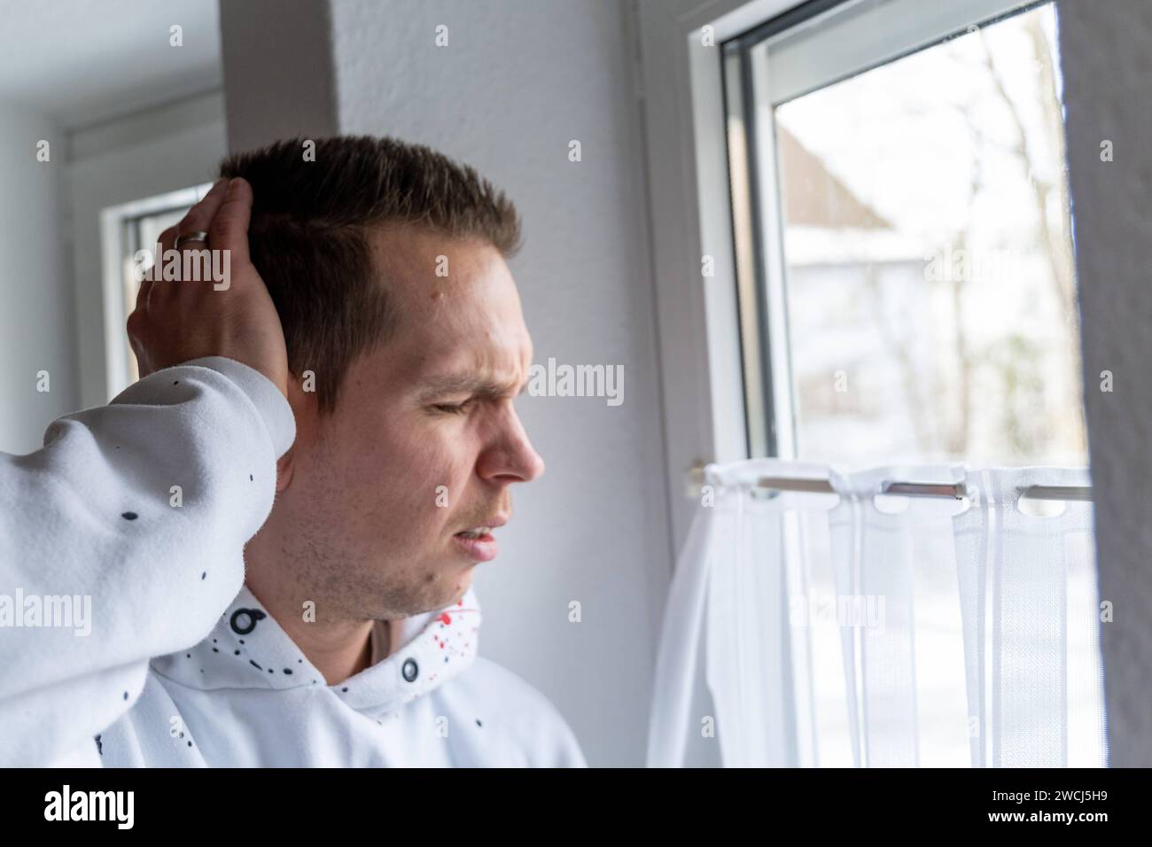 Augusta, Baviera, Germania - 14 gennaio 2024: Un uomo tiene la testa in difficoltà mentre si trova alla finestra. Concetto di mal di testa e malattie *** Ein Mann hält sich den Kopf vor Schmerzen während er am Fenster steht. Kopfschmerzen und Krankheiten Konzept Foto Stock