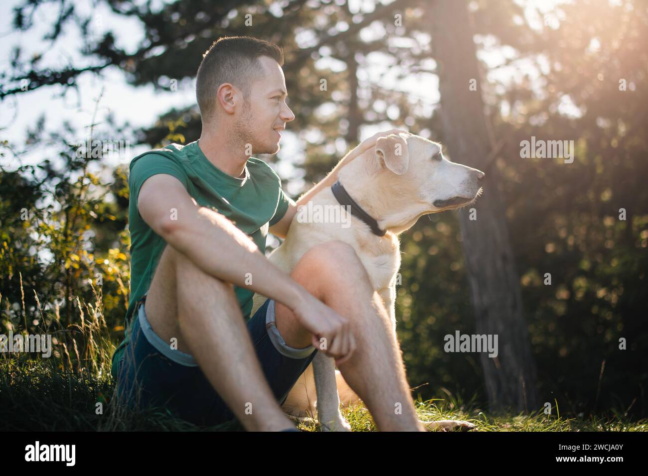 Ritratto di un uomo con un bel cane seduto in erba sotto gli alberi. Felice proprietario del labrador retriever che riposa nella natura nelle soleggiate giornate estive. Foto Stock