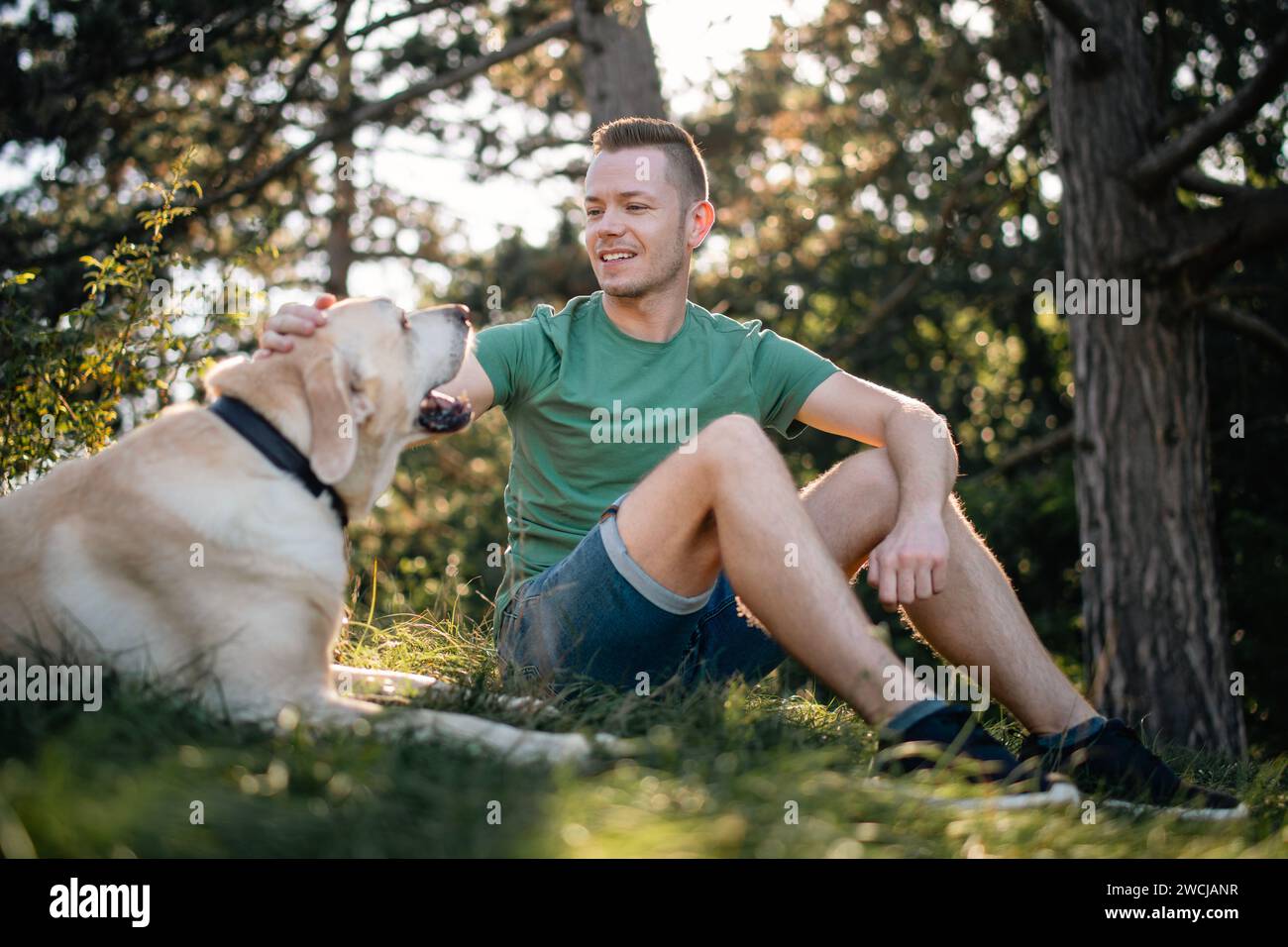 Ritratto di un uomo con un bel cane seduto in erba sotto gli alberi. Felice proprietario del labrador retriever che riposa nella natura nelle soleggiate giornate estive. Foto Stock