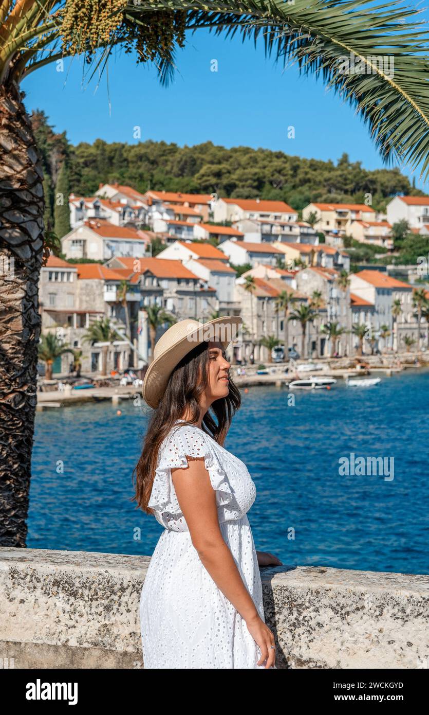 Una donna elegante si erge graziosamente sul mare, trasudando una bellezza senza tempo con il suo vestito bianco fluente e l'elegante cappello di paglia con una città idilliaca come sfondo Foto Stock