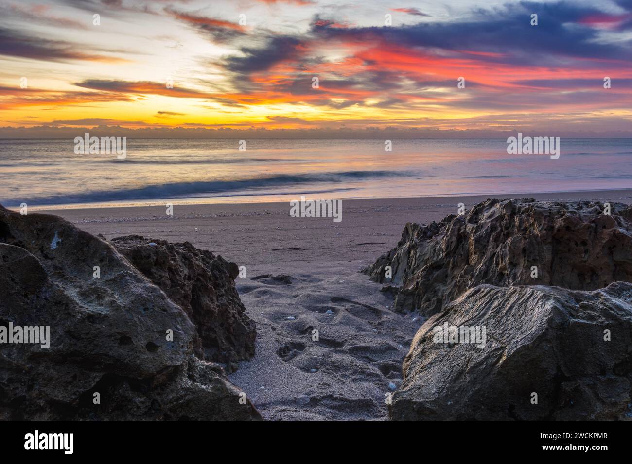 Spettacolare alba sulla spiaggia con nuvole vivaci e terreno roccioso, ideale per contenuti di viaggio vivaci, grafica Web dinamica e sfondi mozzafiato. Foto Stock