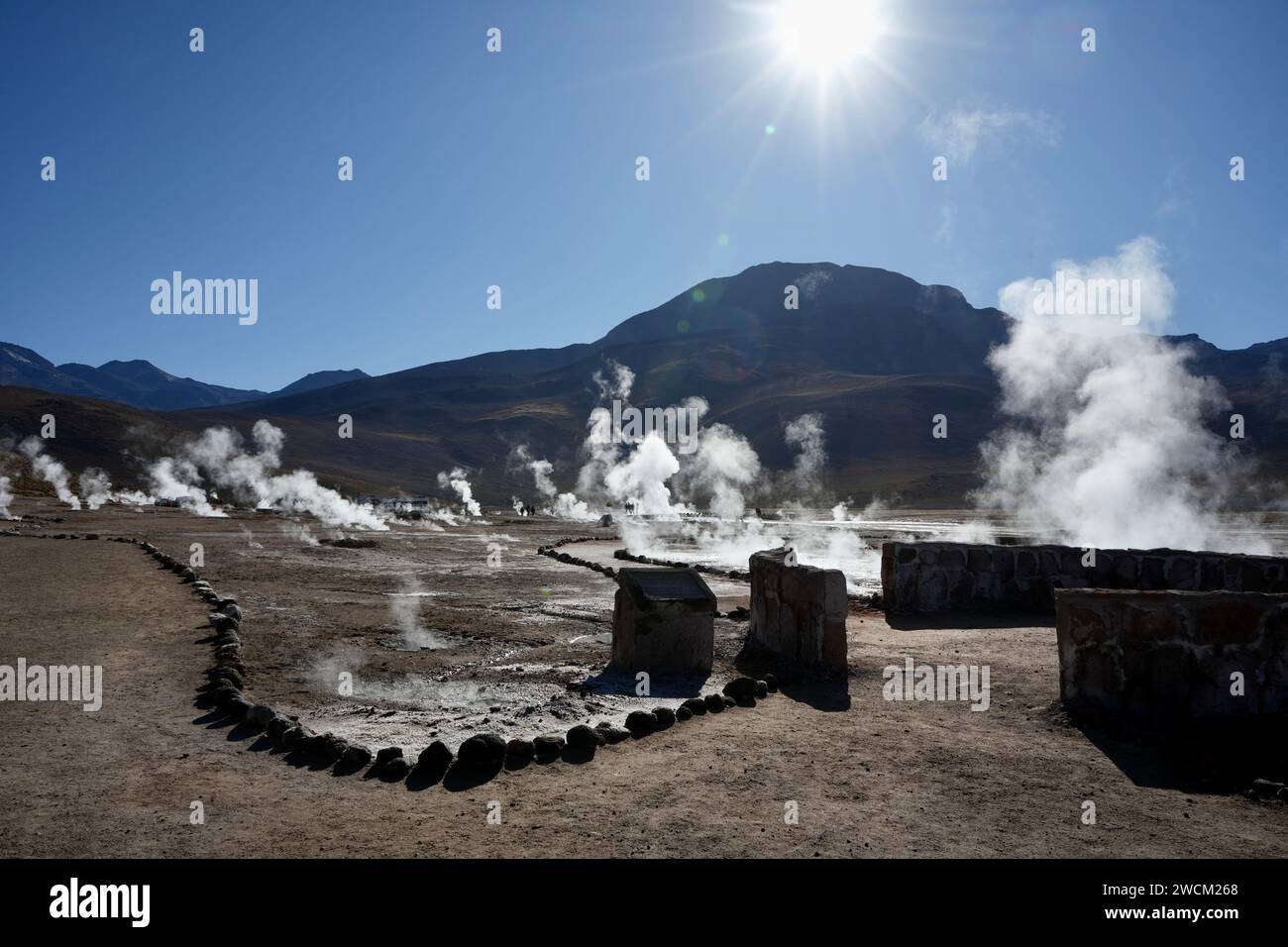 Il vapore si innalza da terra dietro file di pietre con il sole che splende sulle montagne alle spalle. Geysers del Tatio, Antofagasta, Cile. Foto Stock