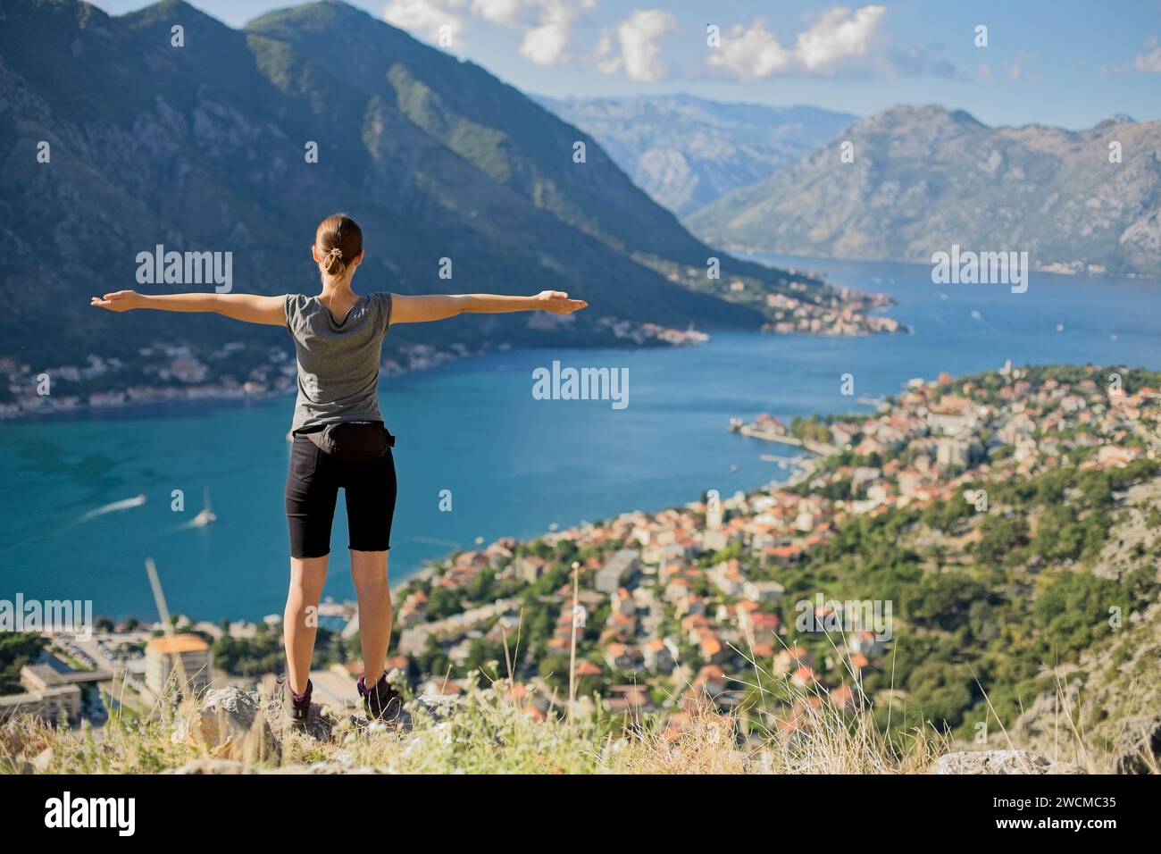Un escursionista si erge a braccia aperte, abbracciando la vista mozzafiato della baia di Cattaro e delle montagne circostanti in Montenegro. Foto Stock