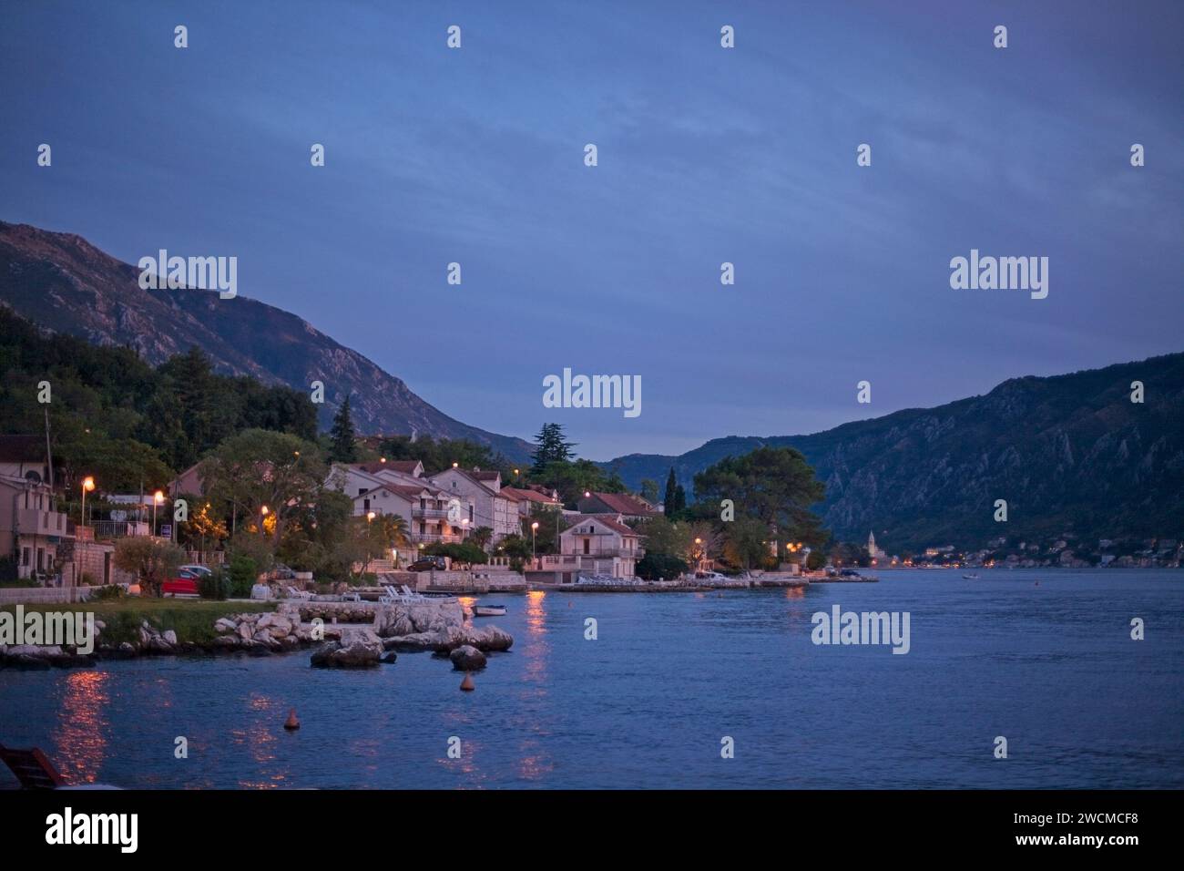 Al calar della sera, il tranquillo villaggio sul mare di Cattaro è illuminato dolcemente, con le calme acque della baia che riflettono l'ultima luce del giorno. Foto Stock