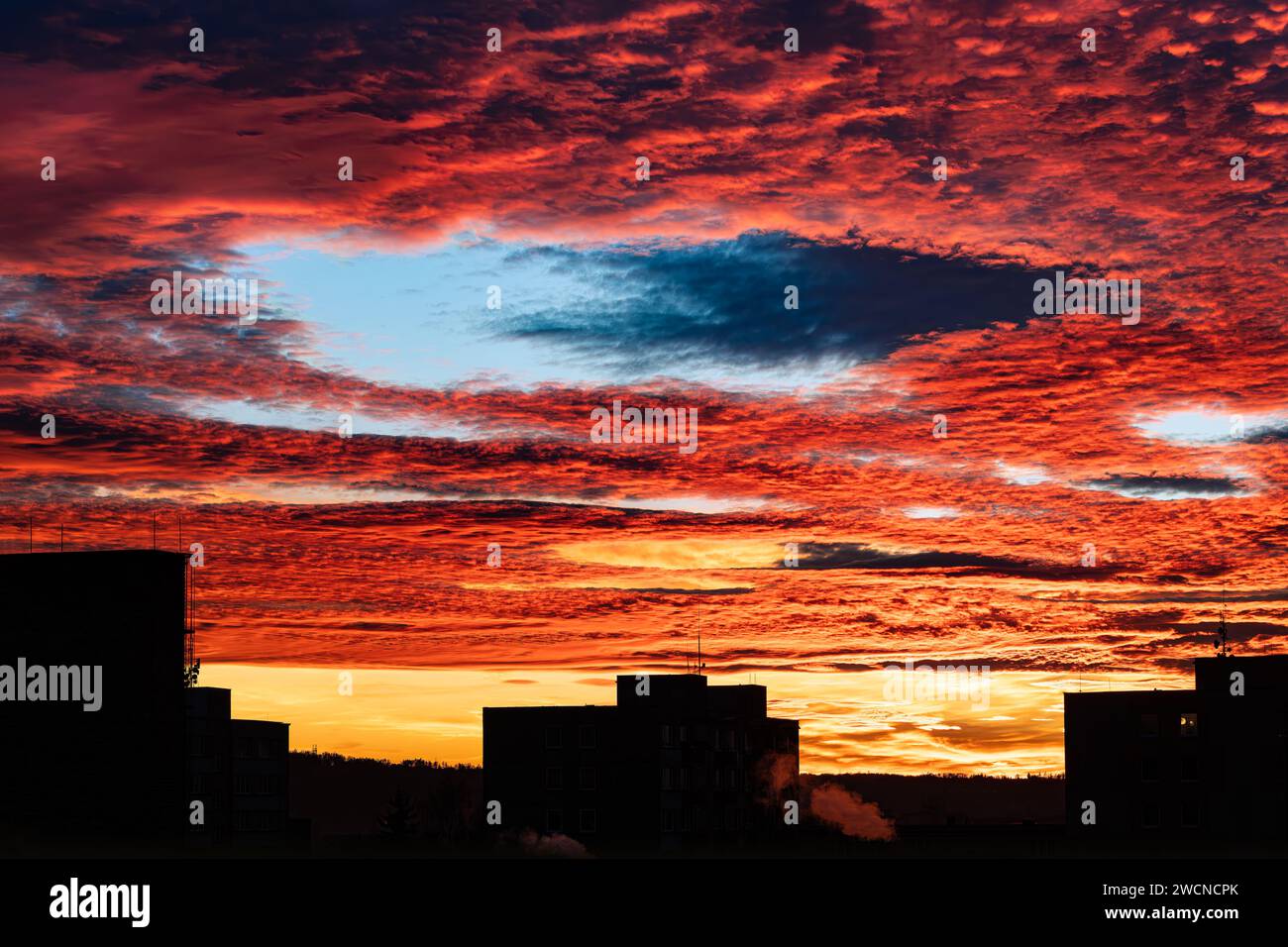 Splendido tramonto con buco a strisce di fallstreak nel cielo rosso ardente e una silhouette di edifici. Maestose nuvole arancioni con grande divario blu al tramonto panoramico d'inverno. Foto Stock