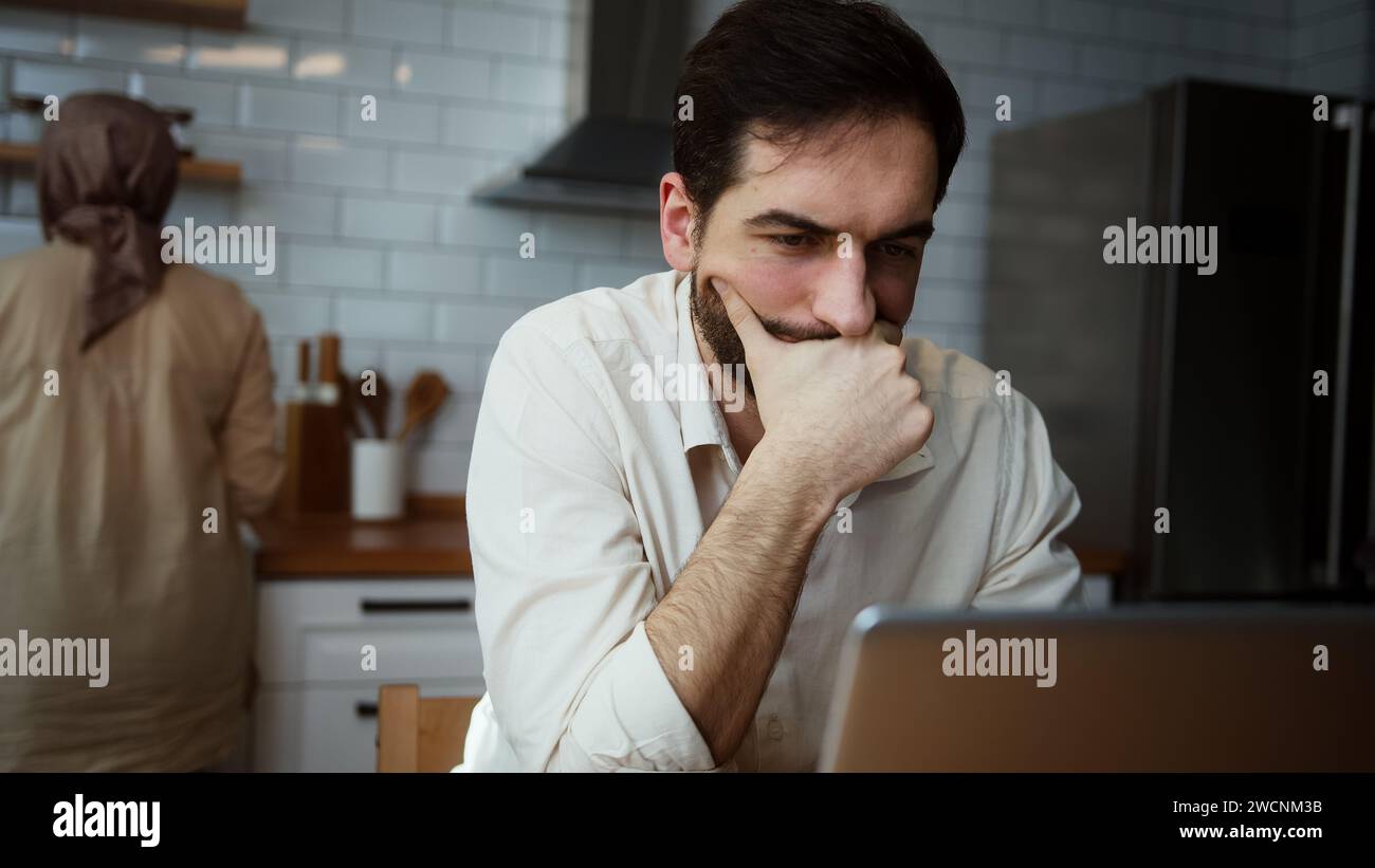 Il budget del marito serio online paga, bollette e finanze calcolano, pagamenti ipotecari utilizzando un computer portatile mentre sua moglie lavora in cucina Foto Stock