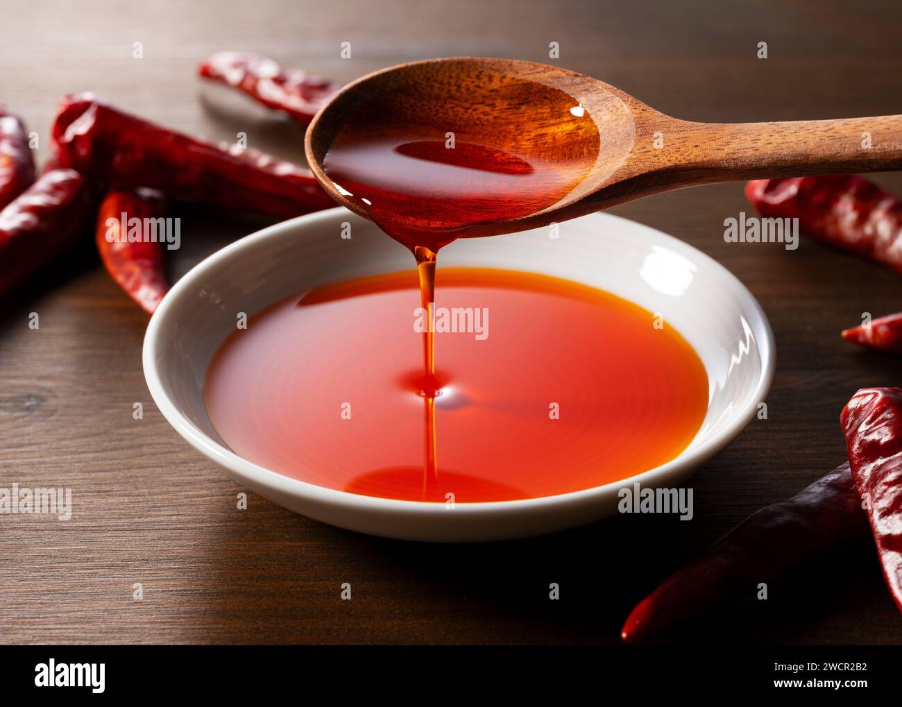 Versare l'olio di peperoncino con un cucchiaio in un piatto di ceramica posto su uno sfondo di legno. Foto Stock