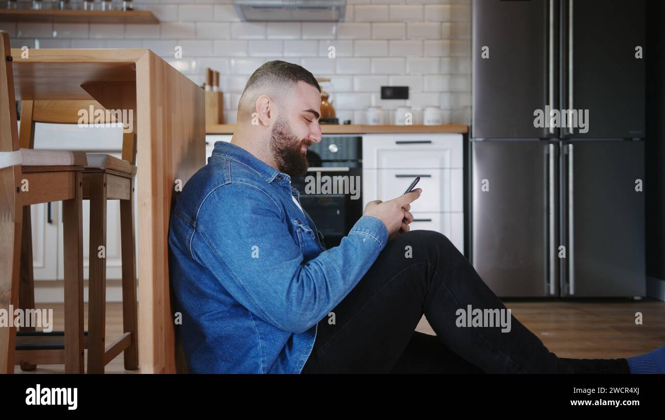 Un giovane adulto barbuto seduto sul pavimento appoggiato contro il bancone della cucina, che scriveva con amici e familiari sul suo smartphone Foto Stock