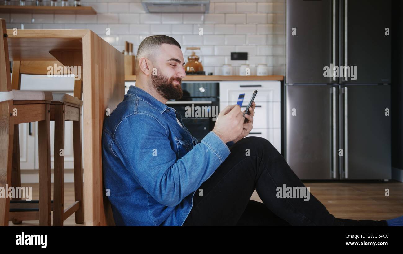 Un giovane adulto barbuto seduto sul pavimento appoggiato contro il bancone della cucina che effettua acquisti con uno smartphone con una carta di credito Foto Stock