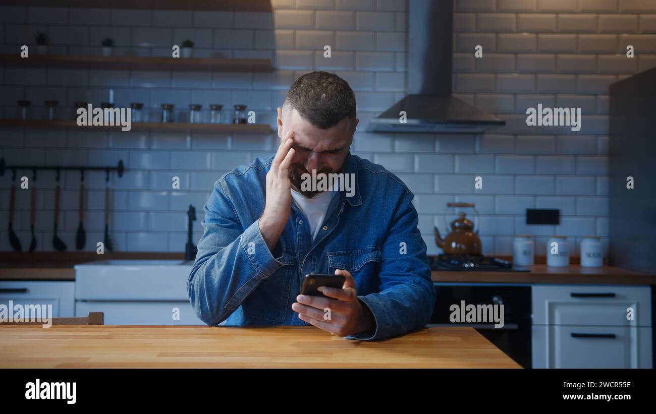 Un giovane adulto barbuto seduto contro il bancone della cucina che ha mal di testa mentre si usa lo smartphone. Un giovane stanco soffre di mal di testa o.. Foto Stock