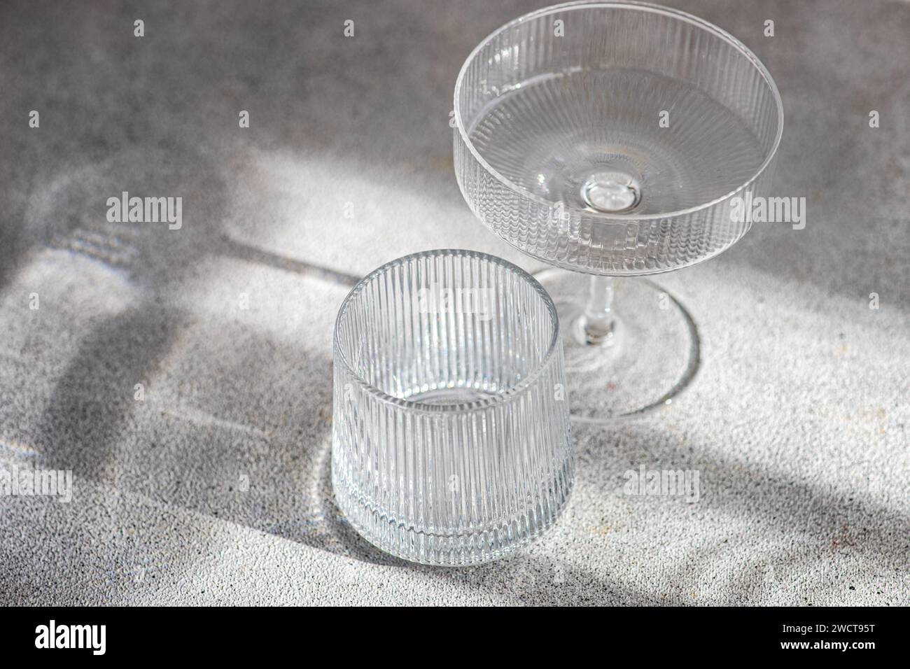 Un bicchiere da cocktail cristallino e un bicchiere sono posizionati su una superficie ruvida, con le loro ombre distinte emesse da una luce soffusa e naturale Foto Stock