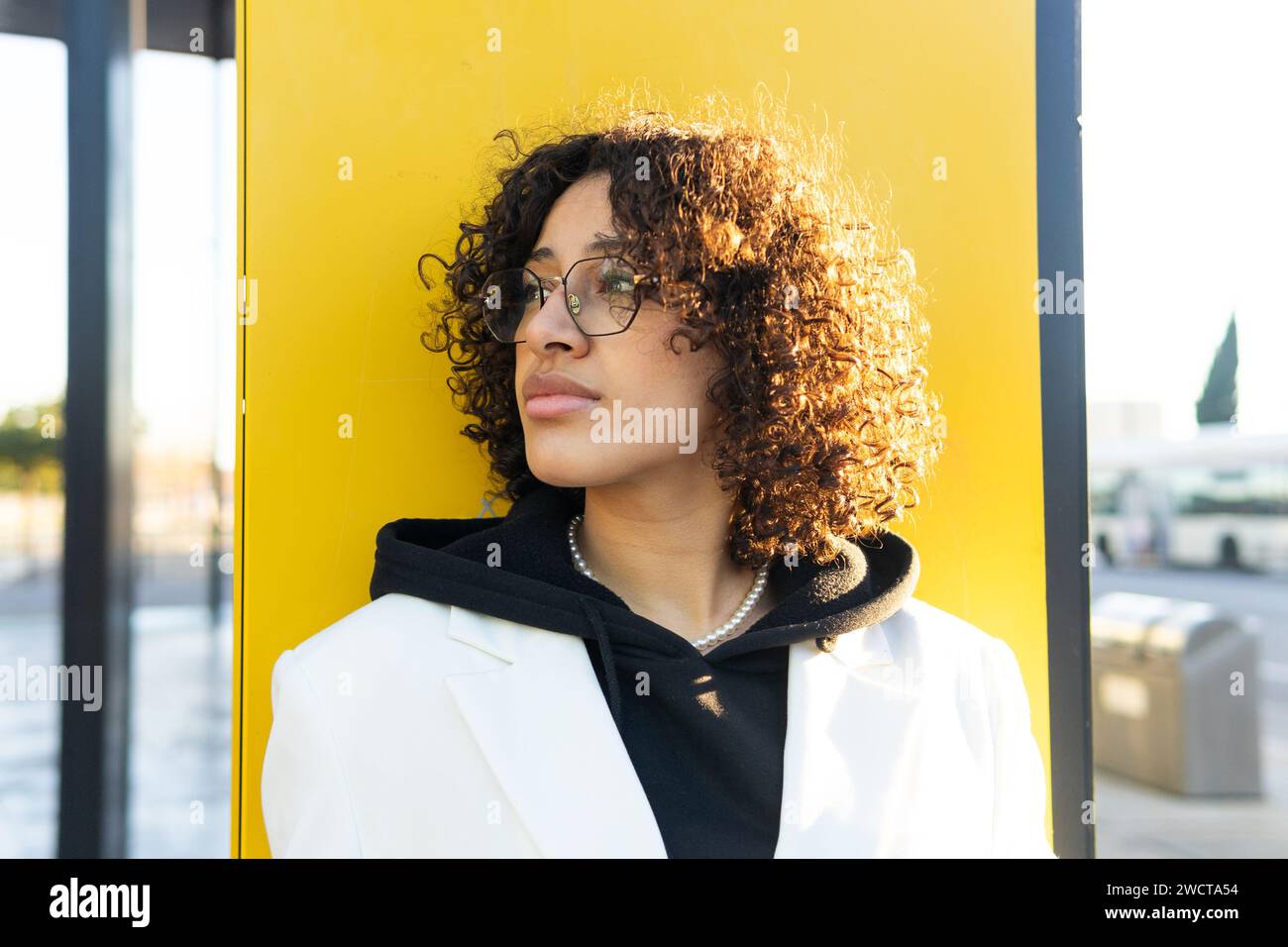 Una giovane donna premurosa con un cappotto bianco e occhiali si appoggia su una parete gialla, guardando in lontananza Foto Stock