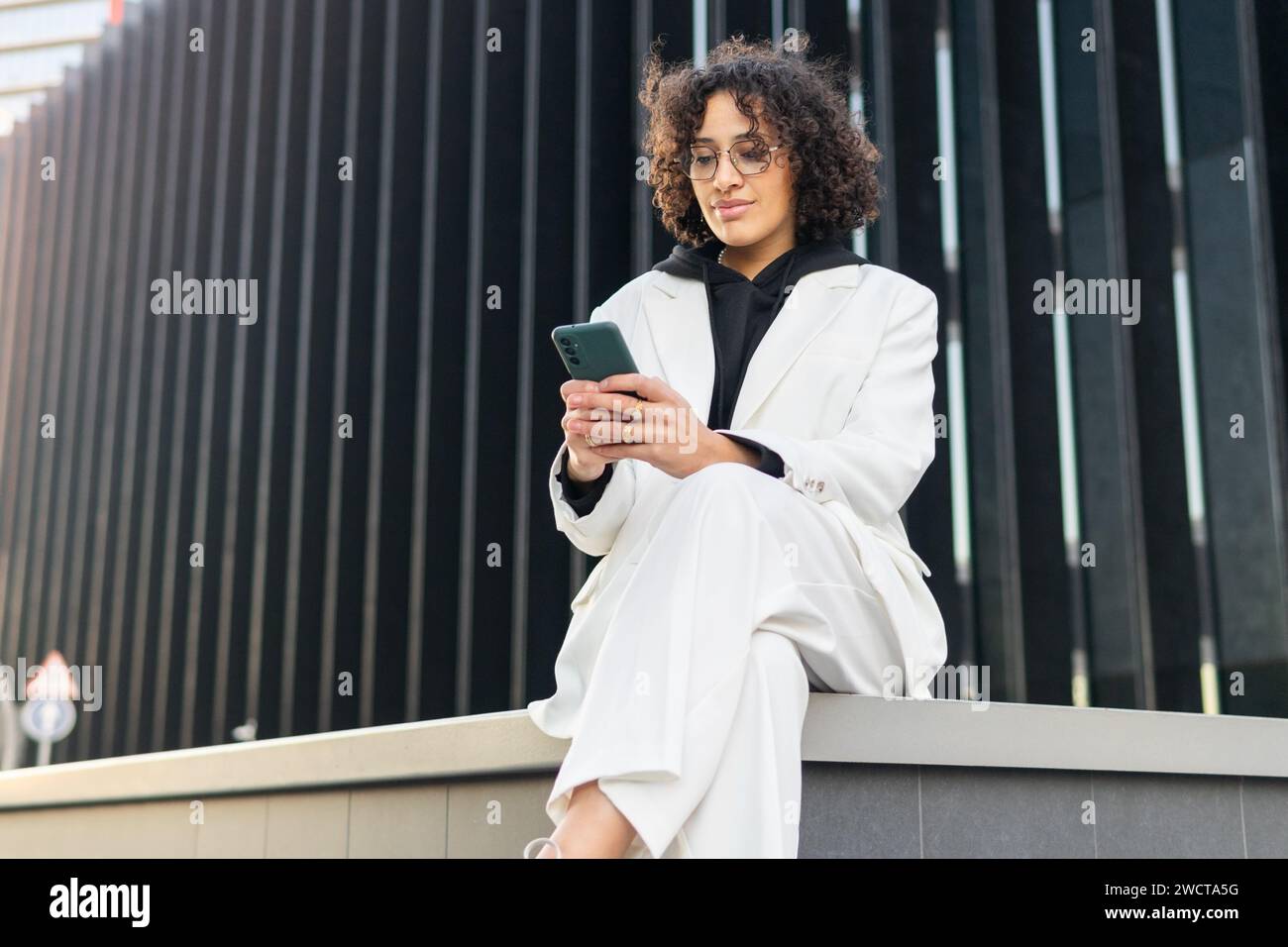 Una giovane donna professionista sicura di sé vestita con un elegante abito bianco si trova all'esterno di un edificio moderno mentre usa lo smartphone Foto Stock
