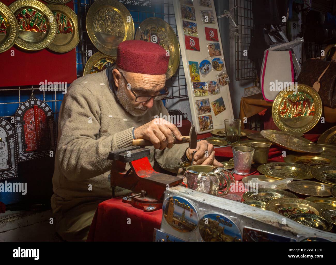 Un artigiano della lavorazione dei metalli che indossa un cappello fez usa un martello presso la sua bancarella turistica di souvenir all'interno del souk nell'antica medina di Sousse, in Tunisia. La m Foto Stock