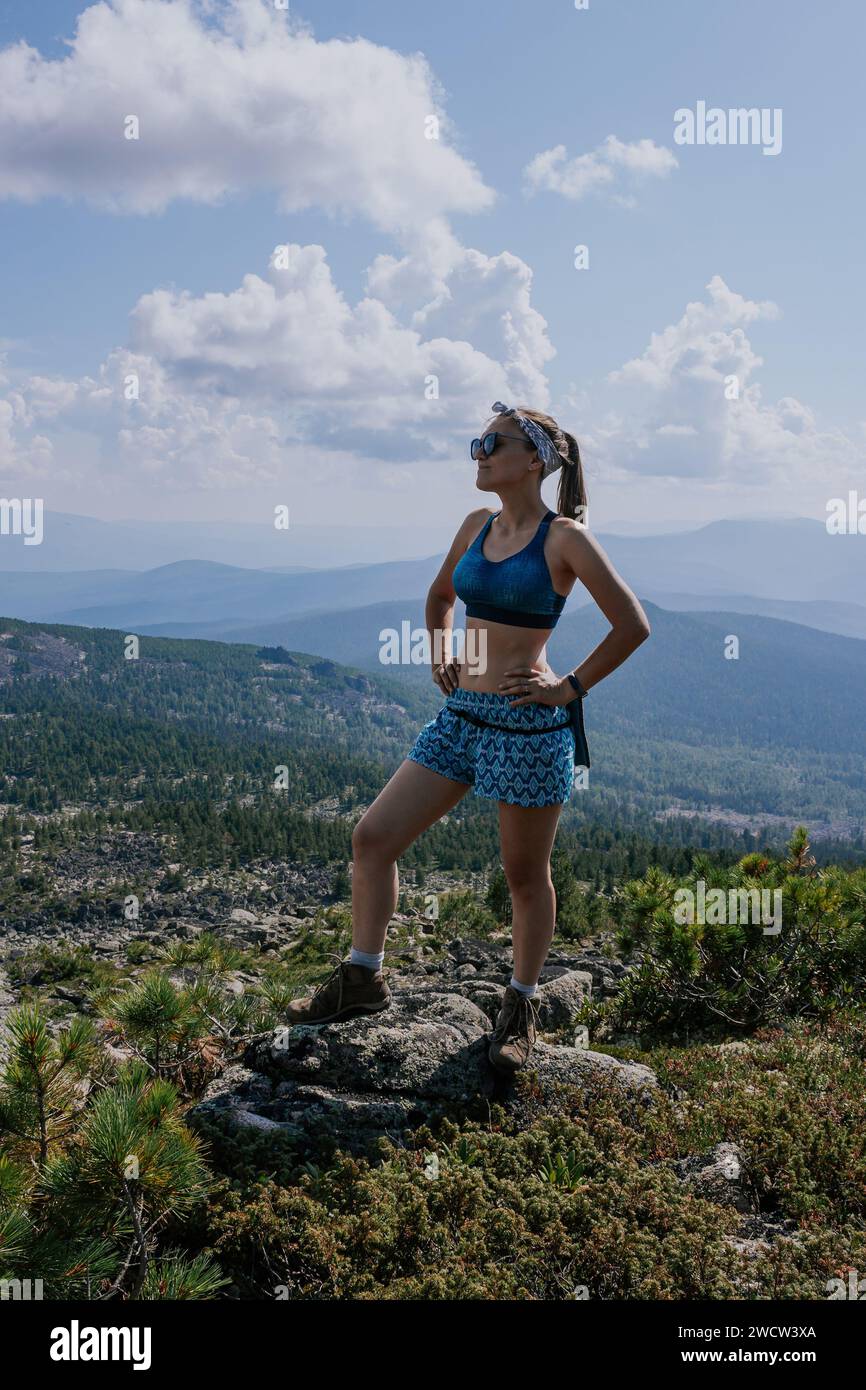 Donna turistica con occhiali da sole, pantaloncini blu e reggiseno sportivo si erge su una pietra sullo sfondo di un paesaggio estivo con verdi colline con conifere Foto Stock