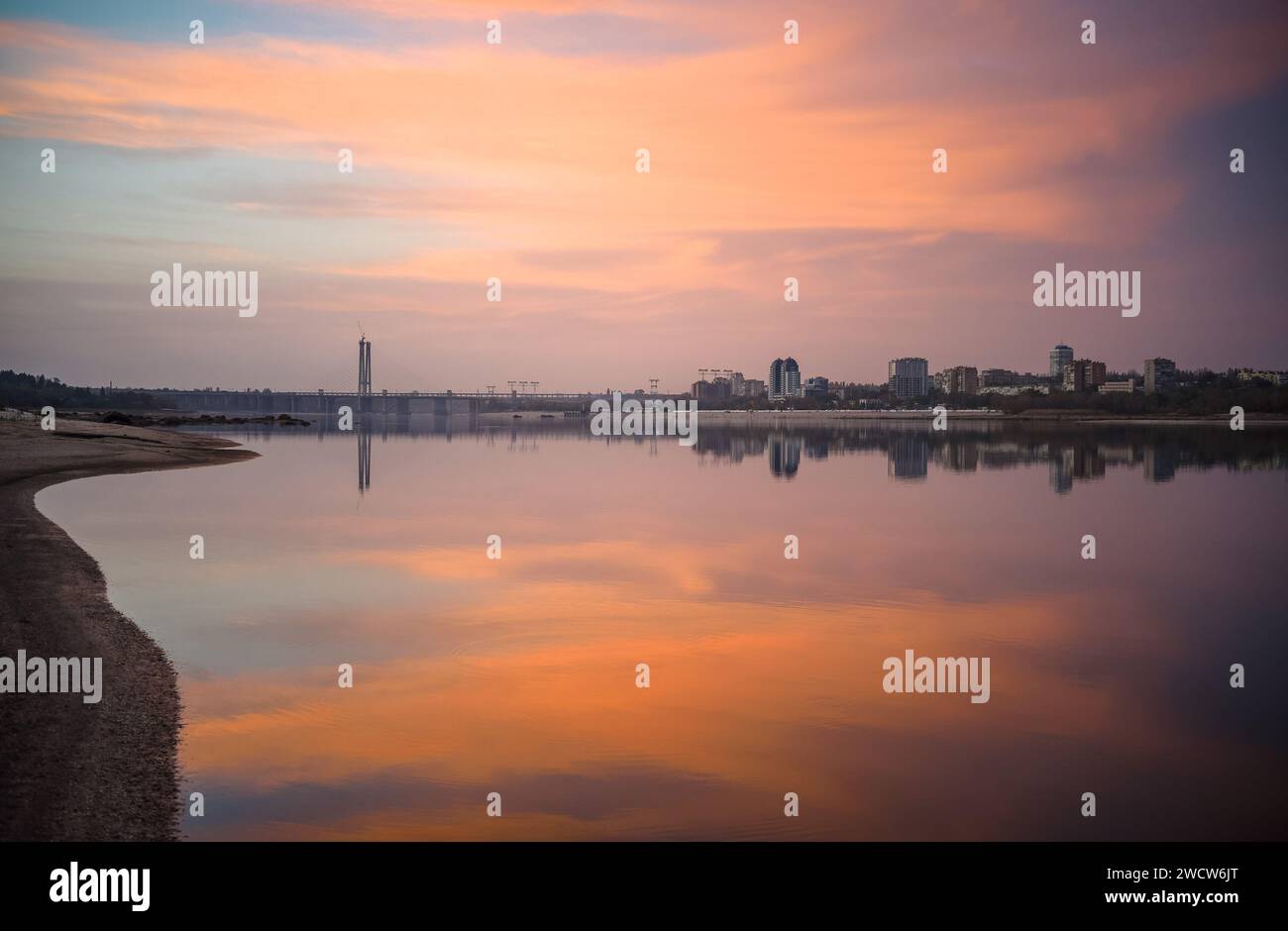 Paesaggio urbano durante il tramonto sulla riva di un ampio fiume con un ponte strallato e alti edifici sulla riva opposta Foto Stock Paesaggio urbano durante il tramonto sulla riva di un ampio fiume con un ponte strallato e alti edifici sulla riva opposta Foto Stock