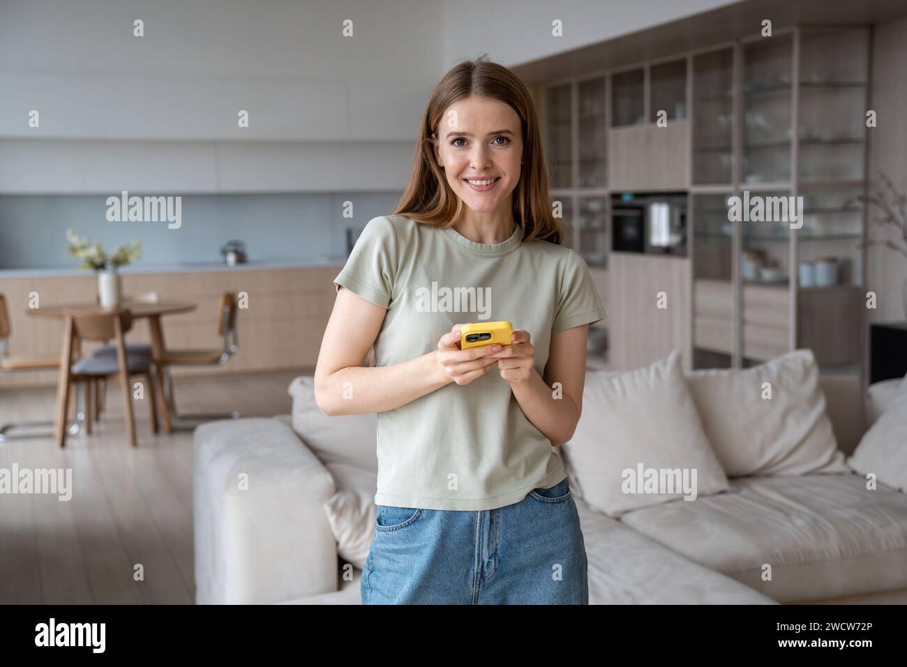 Ritratto sorridente ha soddisfatto la giovane donna di buon umore guardando la fotocamera che tiene lo smartphone a casa. Foto Stock