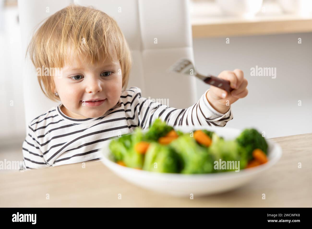 Una bambina carina sta mangiando broccoli e verdure di carote con una forchetta. Foto Stock