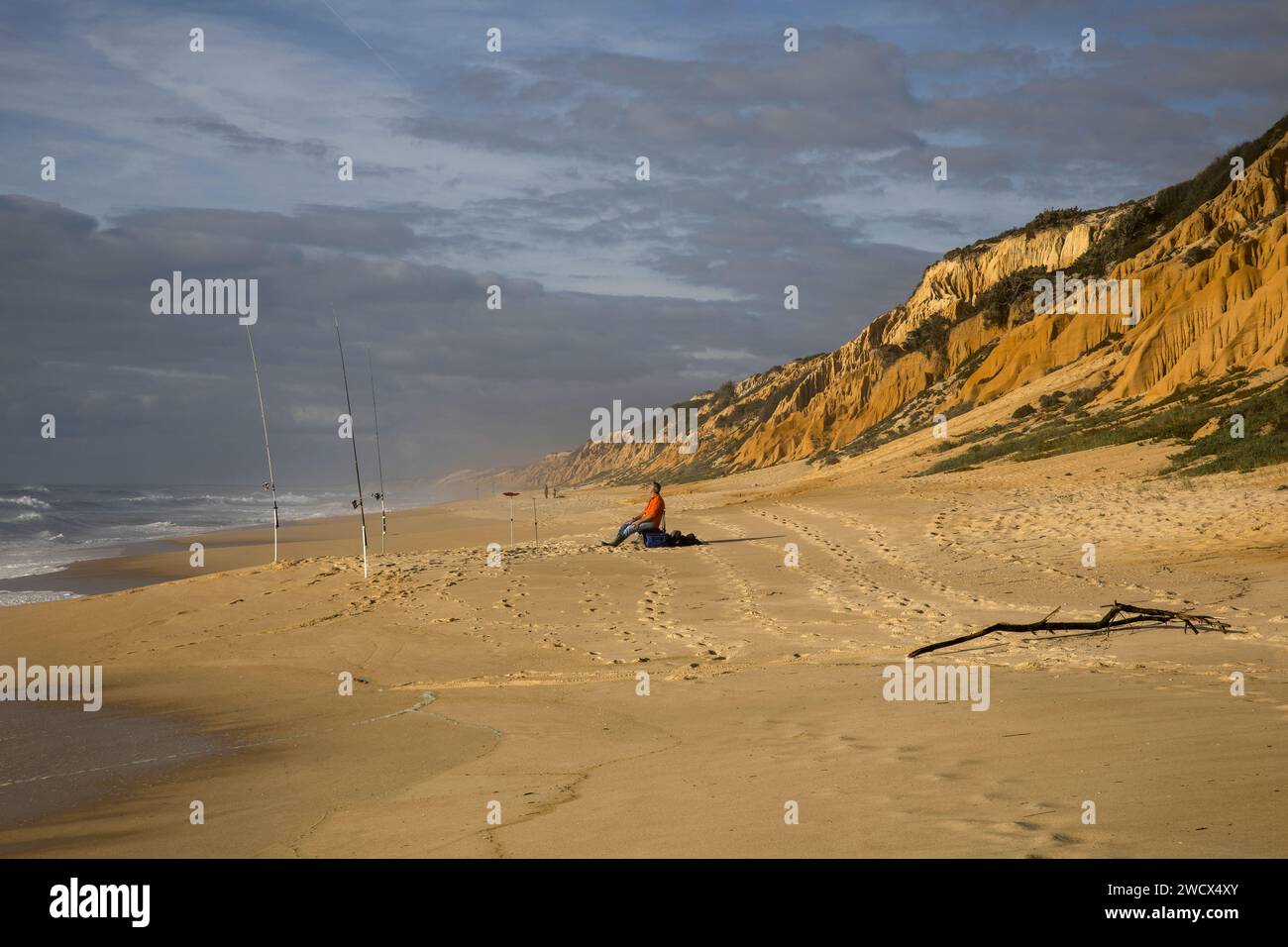 Portogallo, Alentejo, spiaggia di Gale Fontainhas, pescatore seduto sulla spiaggia dove le sue canne da pesca sono piantate tra l'Oceano Atlantico e scogliere fossili ocra risalenti a cinque milioni di anni fa Foto Stock