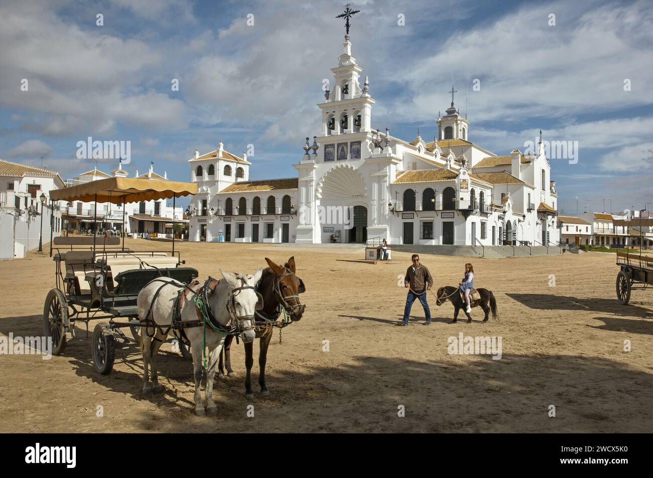 Spagna, Andalusia, El Rocío, uomo che cammina la figlia su un pony in una strada sabbiosa con carri trainati da cavalli di fronte all'hermitage Nuestra señora del Rocío Foto Stock