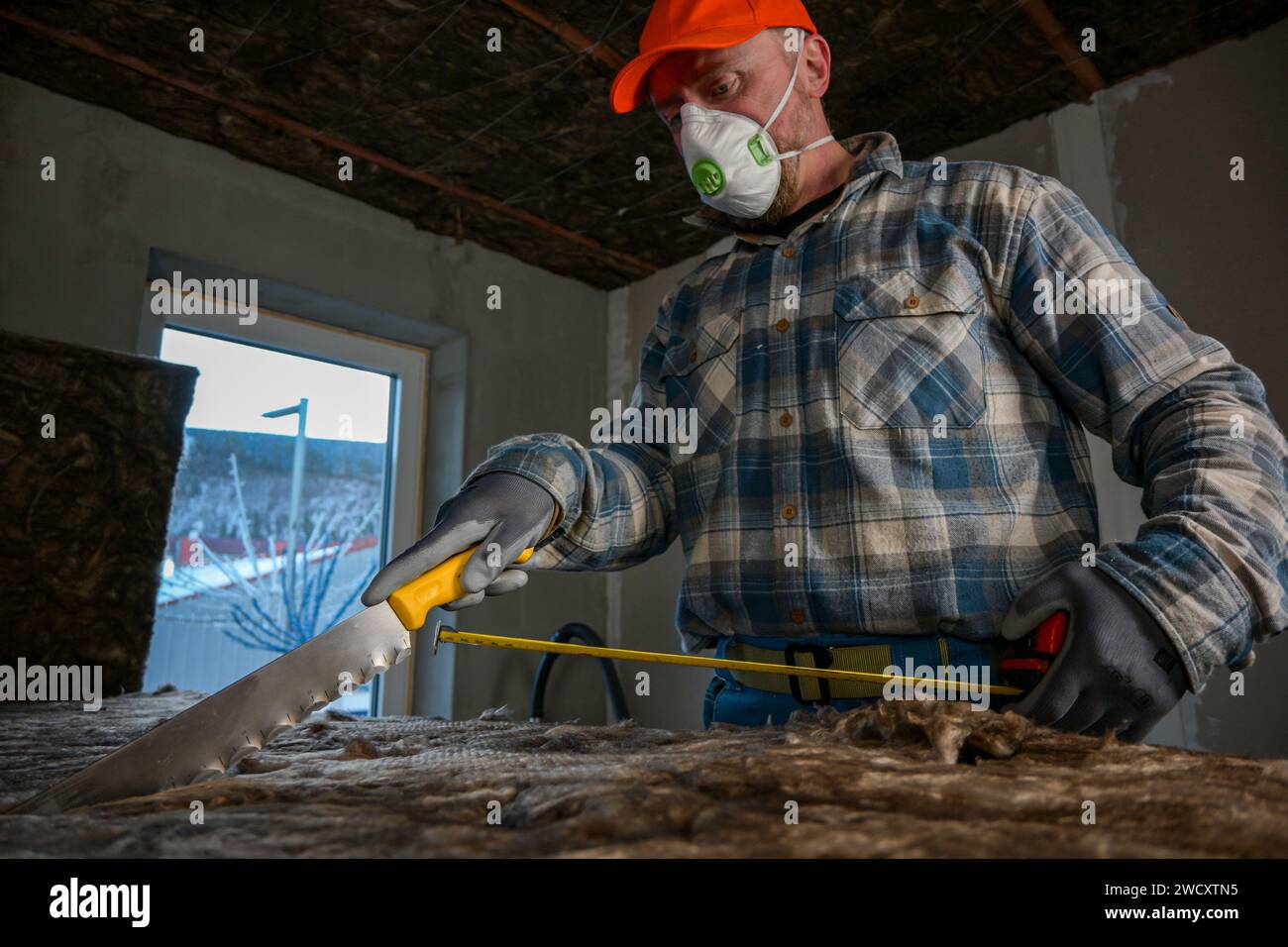 un lavoratore con tute e guanti misura la lunghezza della lana di vetro con un metro a nastro fissato al coltello sulla lana di vetro. Foto Stock