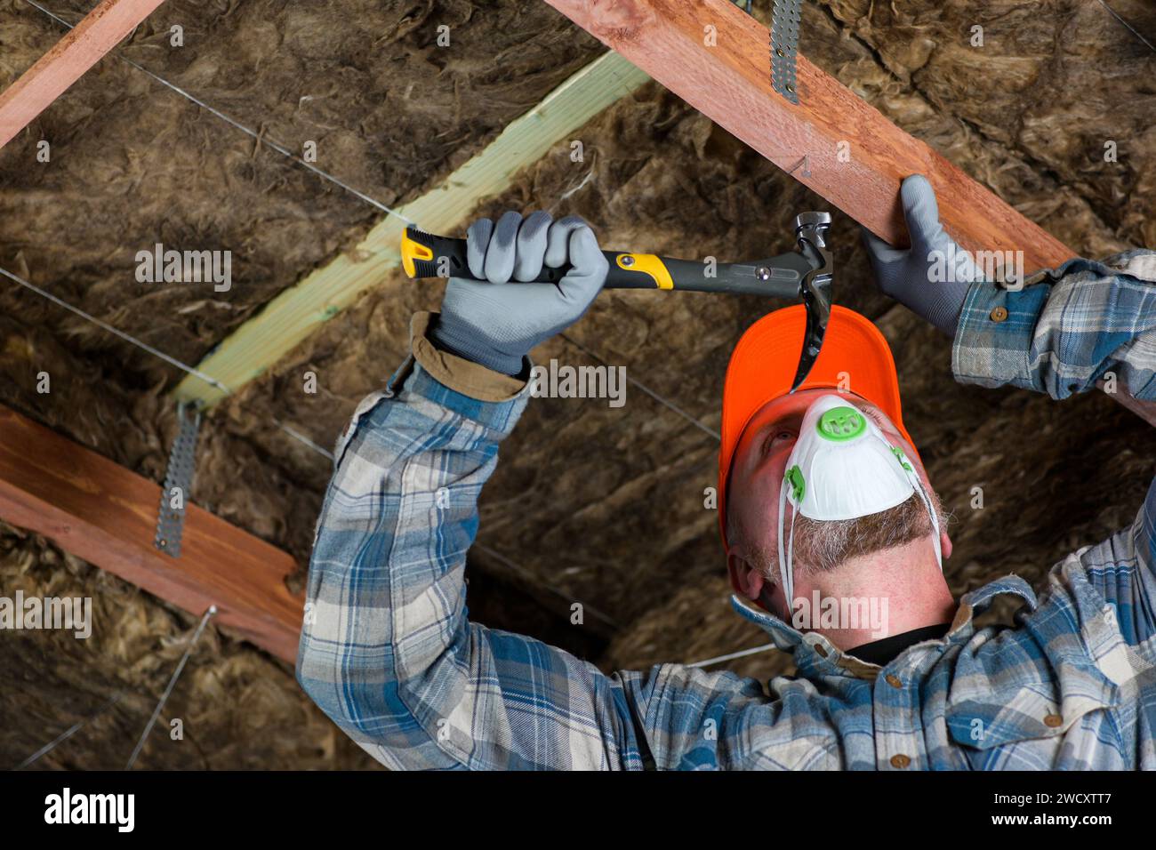 un lavoratore che indossa tute, guanti e un respiratore guida un chiodo nel legno con un martello - preparazione per la lana di vetro nel tetto. Foto Stock