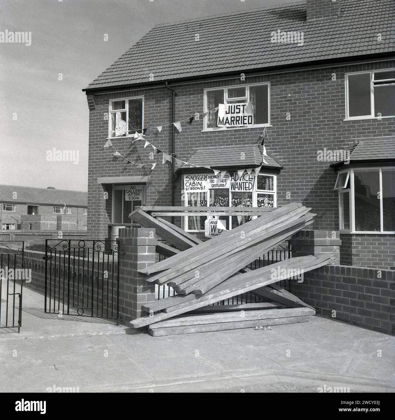 Anni '1960, storica, appena sposata....in una nuova tenuta abitativa, tavole in legno che barricano il vialetto di una casa di recente costruzione decorata con bunting e divertenti avvisi di matrimonio, gli slogan appena sposati includono: "Niente chiamanti dopo le 18:30, cabina degli Snoggers - lezioni!, nuovi Weds - non disturbare, Consiglio ricercato! Foto Stock