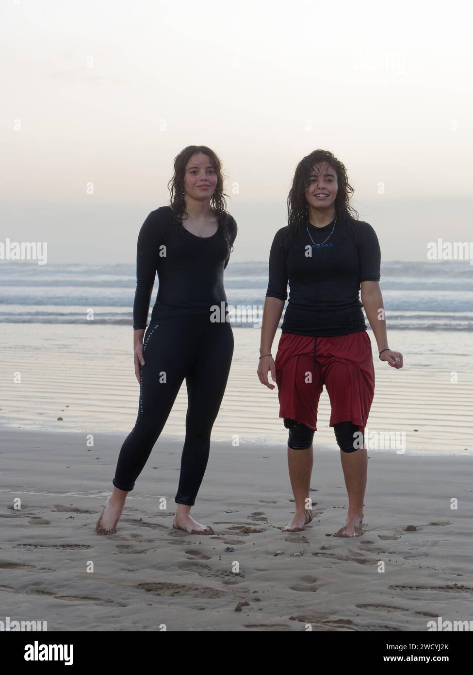 Due giovani donne in costume da bagno e capelli bagnati si stendono sulla spiaggia dopo un tuffo al tramonto in mare a Essaouira, Marocco, 17 gennaio 2024 Foto Stock