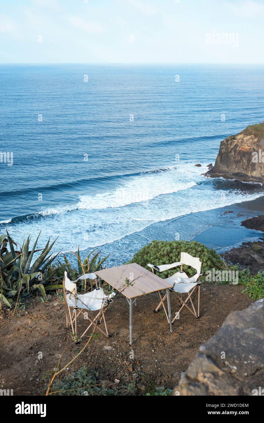 Una scena tranquilla con due sedie da campeggio vuote e un tavolo, invitante collocato sul bordo di una scogliera che si affaccia sull'oceano, catturati dalla luce del giorno Foto Stock
