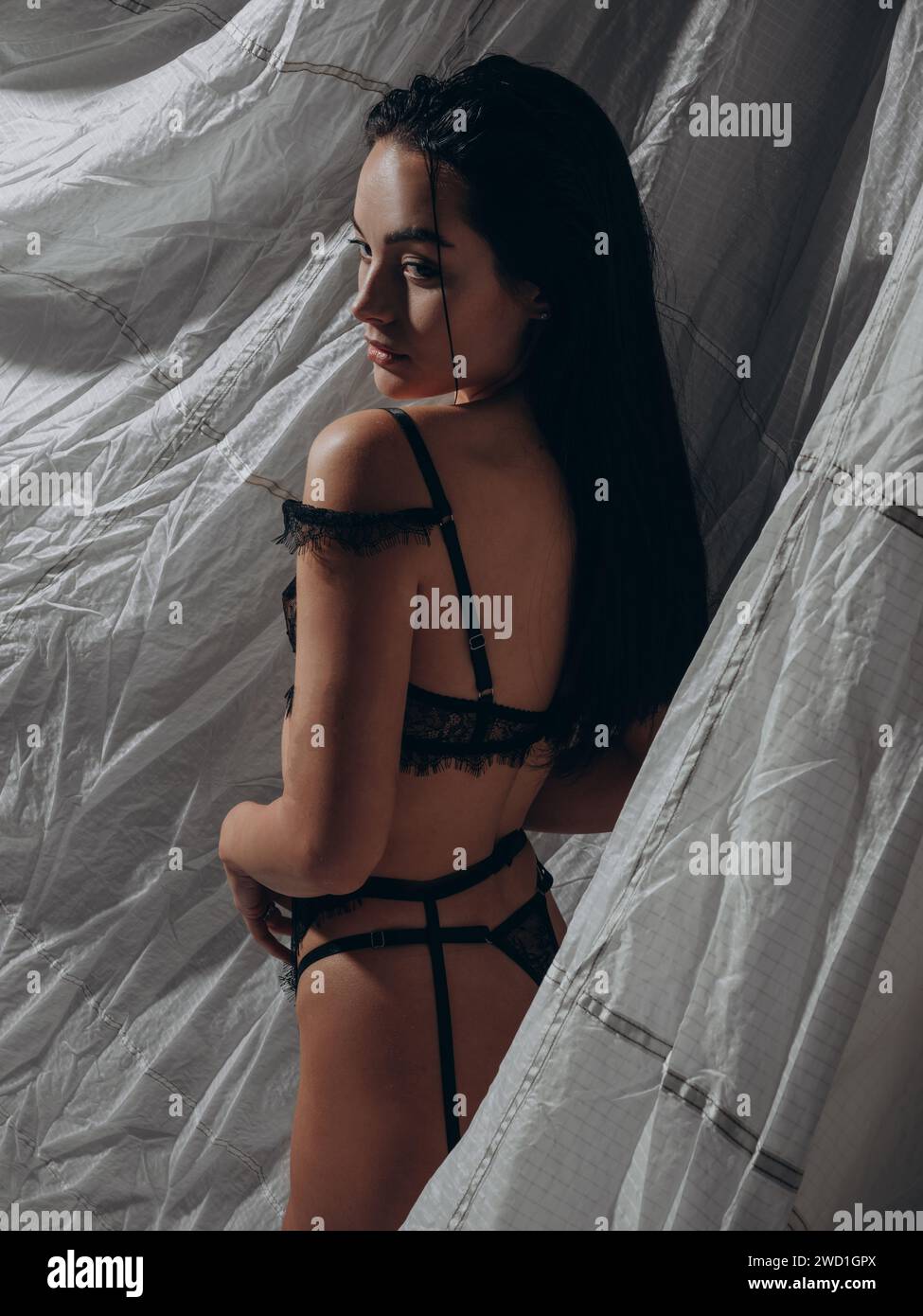 Giovane donna seducente e sexy che posa in lingerie nera lacy e cintura con sospensioni in studio scuro su sfondo grigio. Stile BDSM. Primo piano. Vista posteriore. Foto Stock