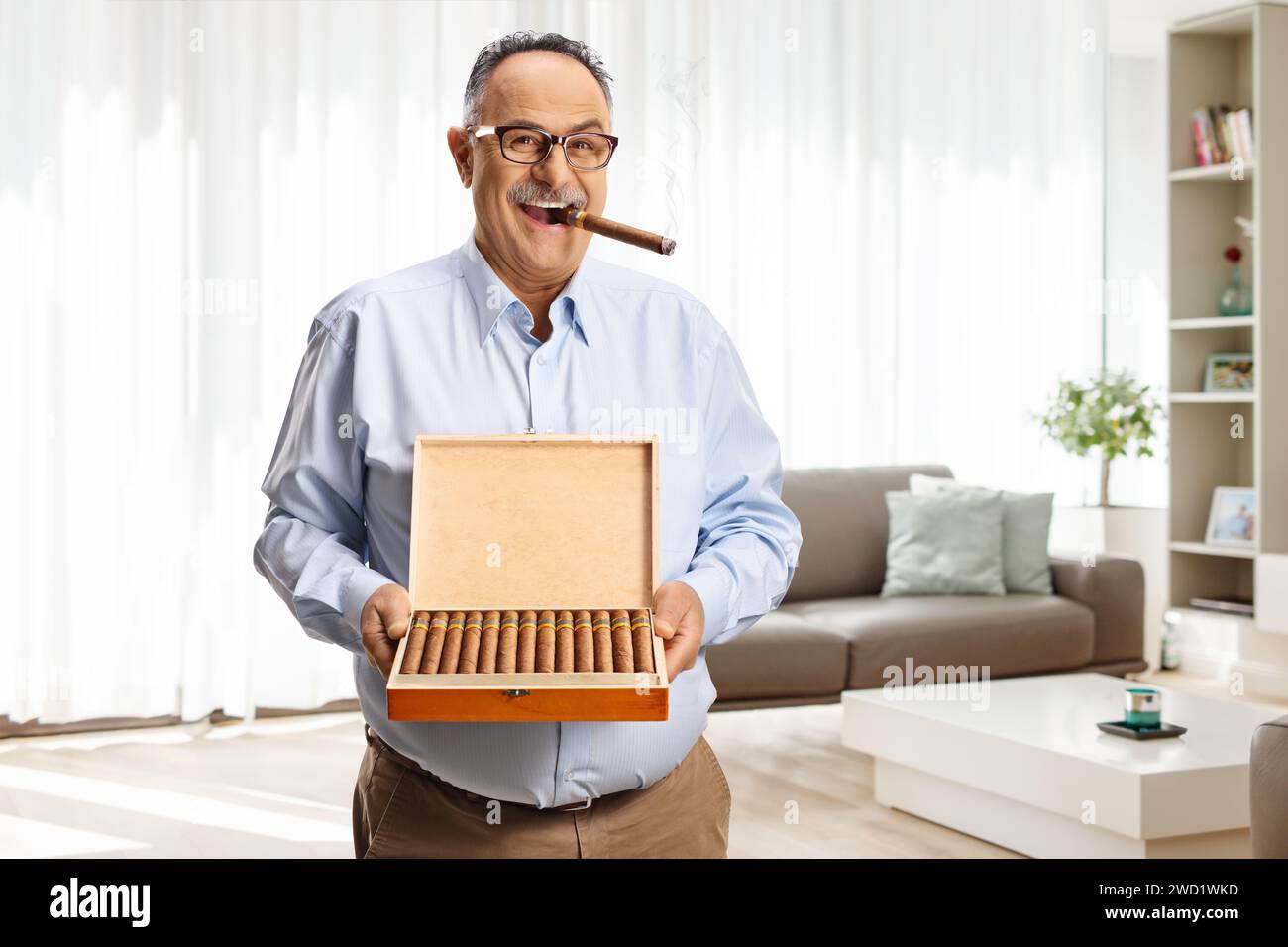 Uomo maturo che fuma un sigaro a casa e tiene una scatola di sigari Foto Stock